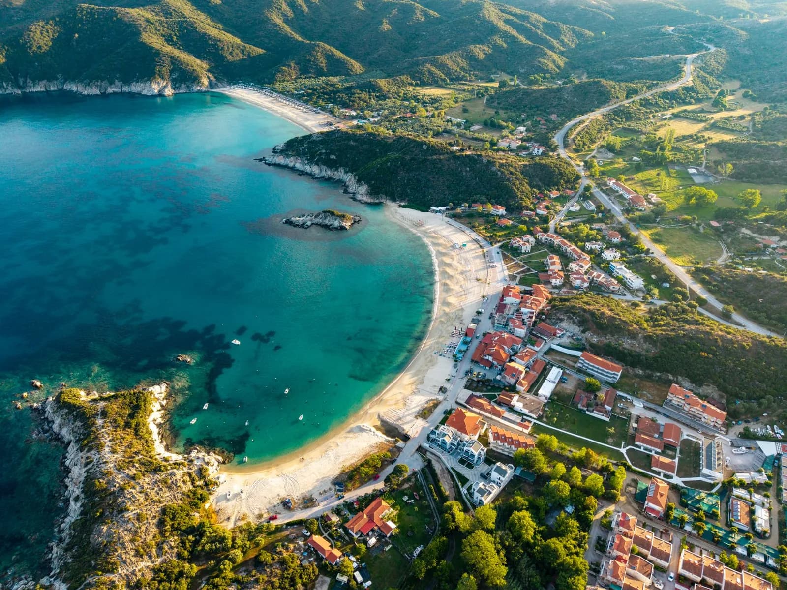 Aerial view of Kalamitsi bay on the southern tip of Sithonia, Halkidiki, a curved sand beach with a small rocky islet in turquoise water and the village's red-roofed houses and green hills behind