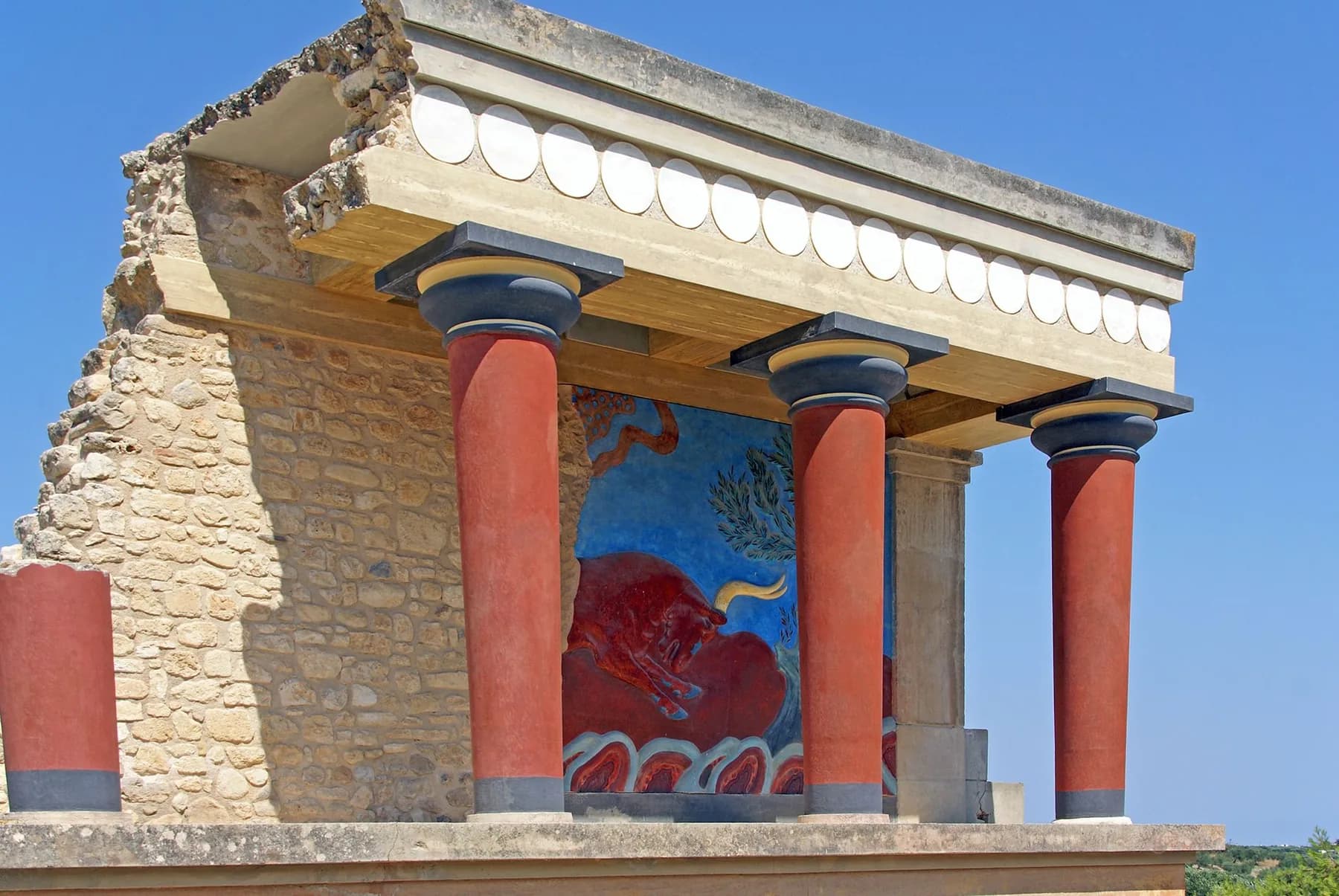 The reconstructed north entrance portico at Knossos, with tapered red columns, a concrete lintel and the charging bull relief set into a blue panel above ancient rubble walling