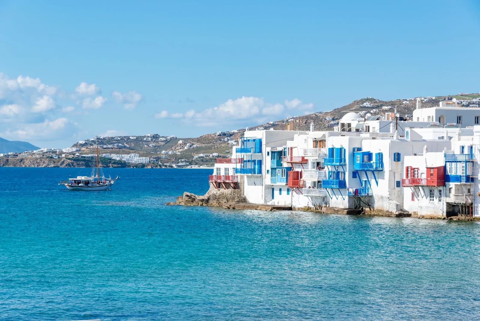 The balconies of Little Venice hanging over the water on the west side of Mykonos Chora, with the hillside of the town behind and a wooden boat offshore