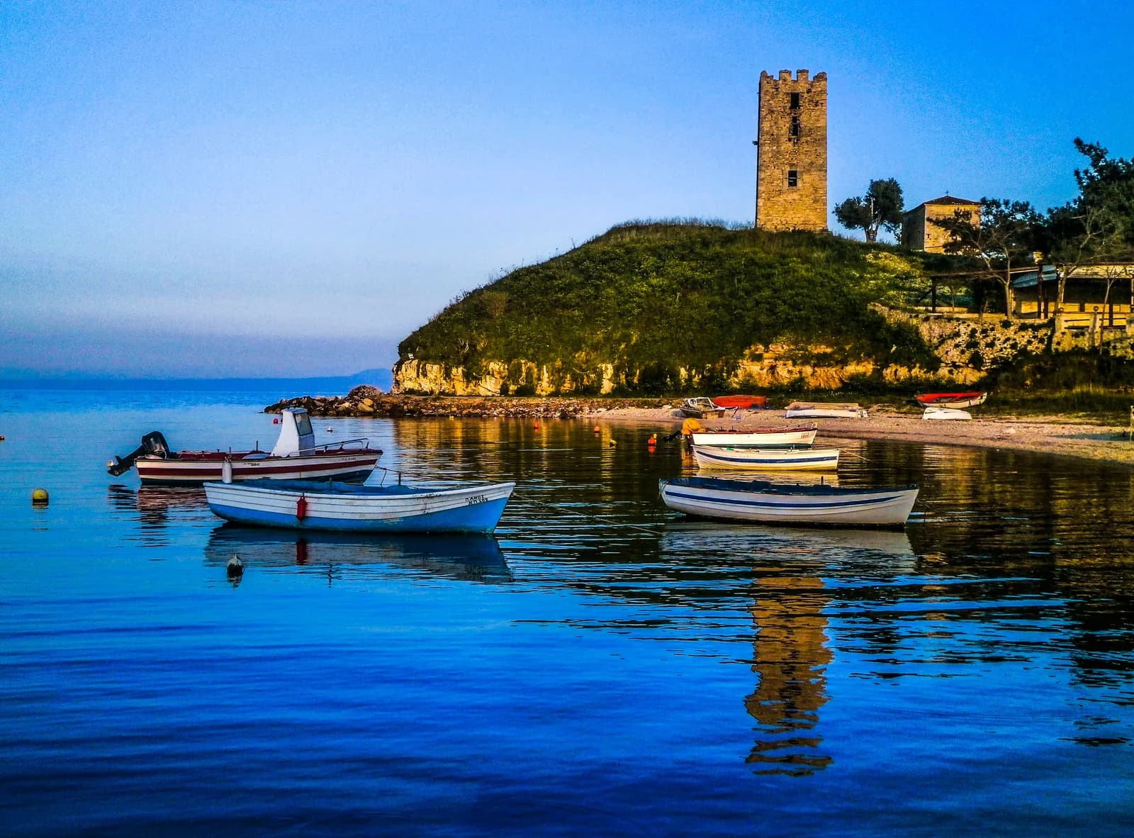 The Byzantine tower of Nea Fokea on its grassy headland above the harbour, with fishing boats moored in calm water, on the Kassandra peninsula of Halkidiki