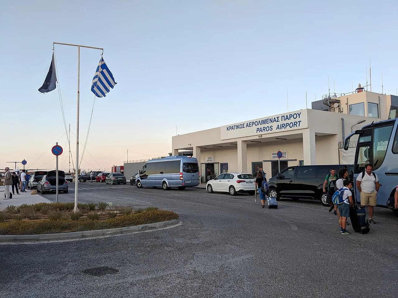 The single-storey terminal of Paros National Airport with its bilingual sign reading Paros Airport, the control tower behind, minibuses and taxis parked outside and passengers with luggage in the forecourt at dusk