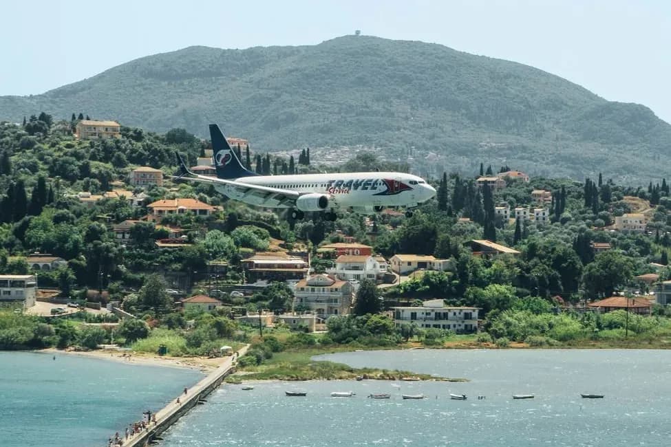 An airliner on final approach to Corfu airport passing low over the Kanoni causeway and the lagoon, with spectators on the footbridge