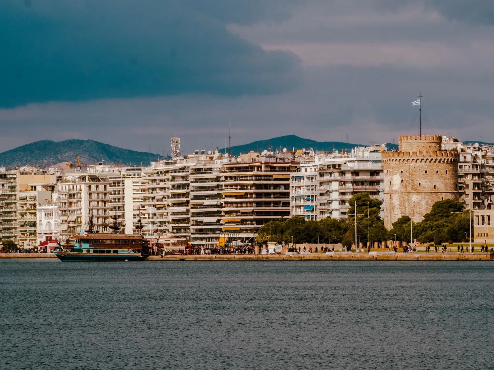 The White Tower and the Nea Paralia seafront of Thessaloniki seen from the Thermaic Gulf, with apartment blocks, a tourist boat and the hills behind the city