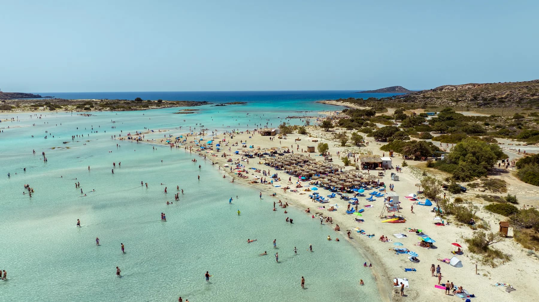 The shallow lagoon at Elafonissi in south-western Crete, with bathers standing in ankle-deep water, rows of straw umbrellas along the sandbar and dry scrub hills behind