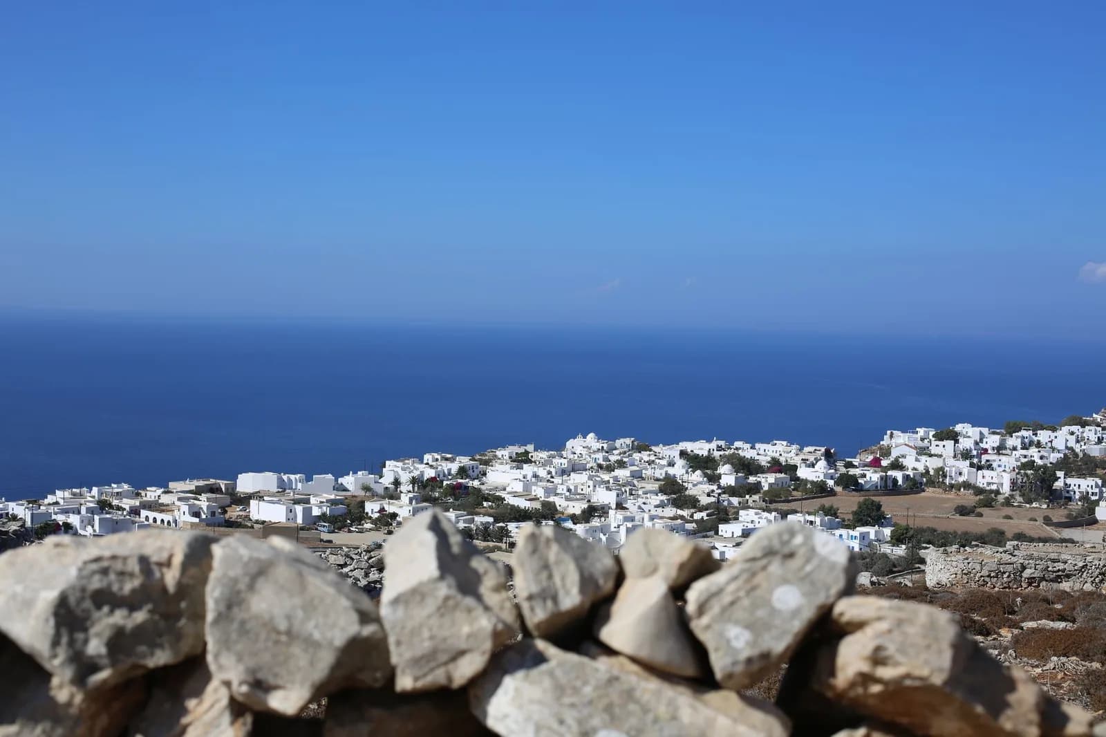 A whitewashed Cycladic village spread down a slope above the Aegean, seen over a dry stone wall