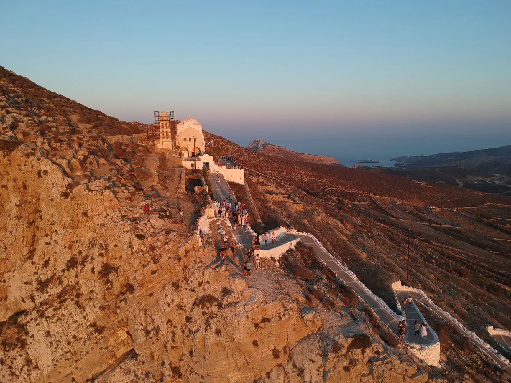 A whitewashed church on a bare ridge above a Cycladic chora at sunset, with a crowd strung out along the zigzag path up to it