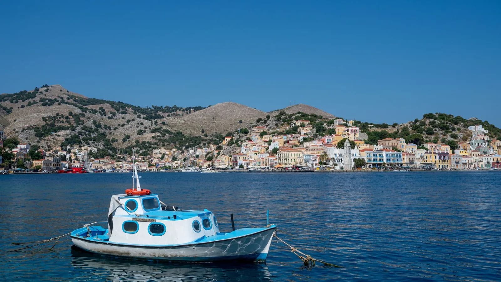 The harbour of Gialos on Symi seen from the water, a small blue and white boat moored in the foreground and the ochre neoclassical houses of the town stacked under bare brown hills behind