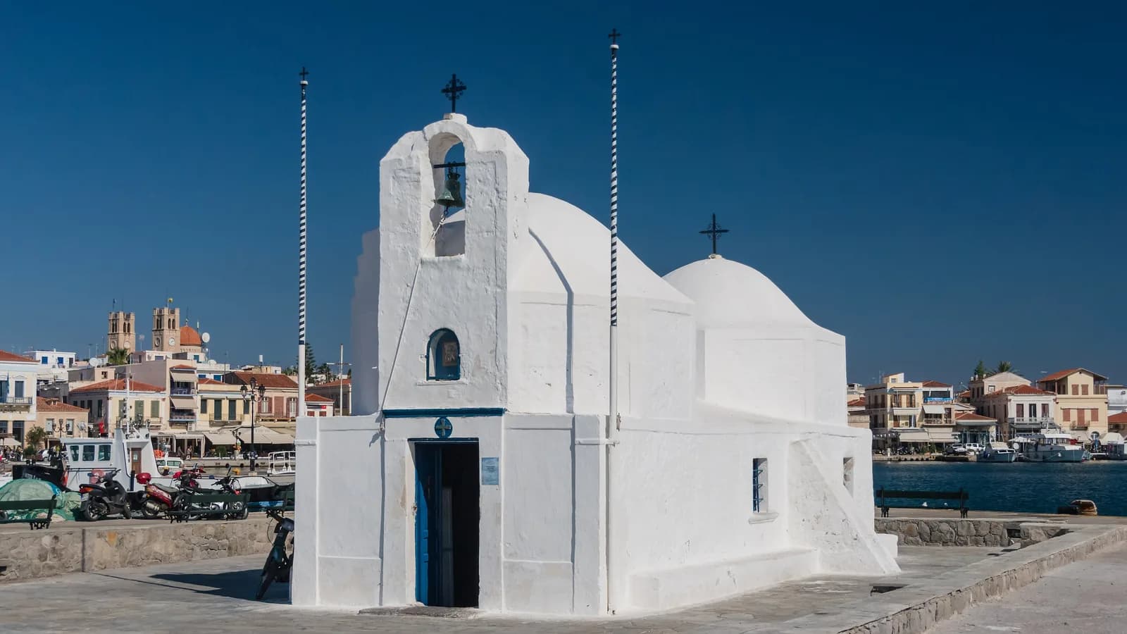 Whitewashed Agios Nikolaos chapel on the harbour quay in Aegina town, with the waterfront and cathedral bell towers behind