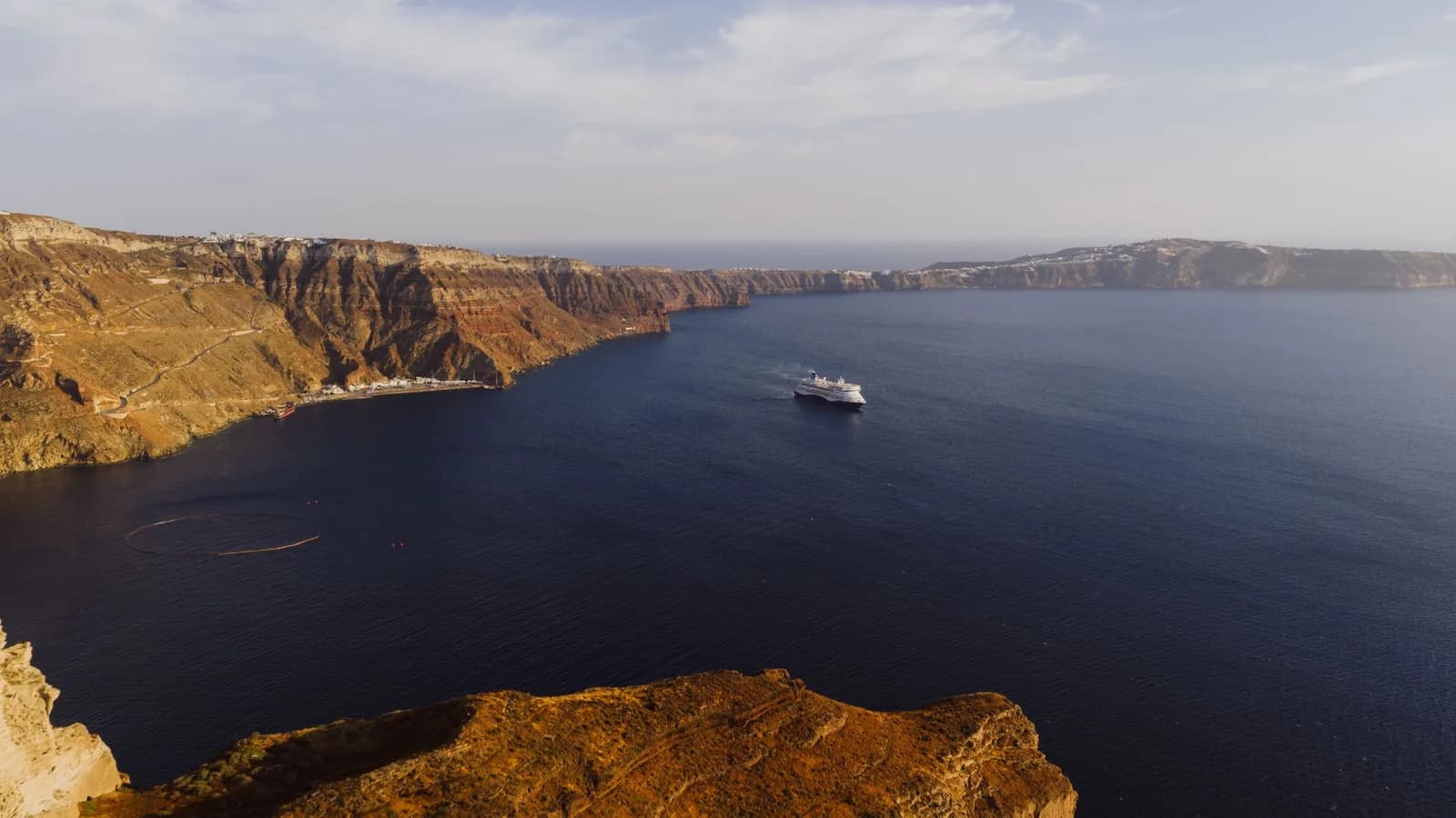 The ferry quay of Athinios on Santorini seen from the caldera rim, a narrow concrete shelf at the base of the cliff with a switchback road climbing the crater wall above it