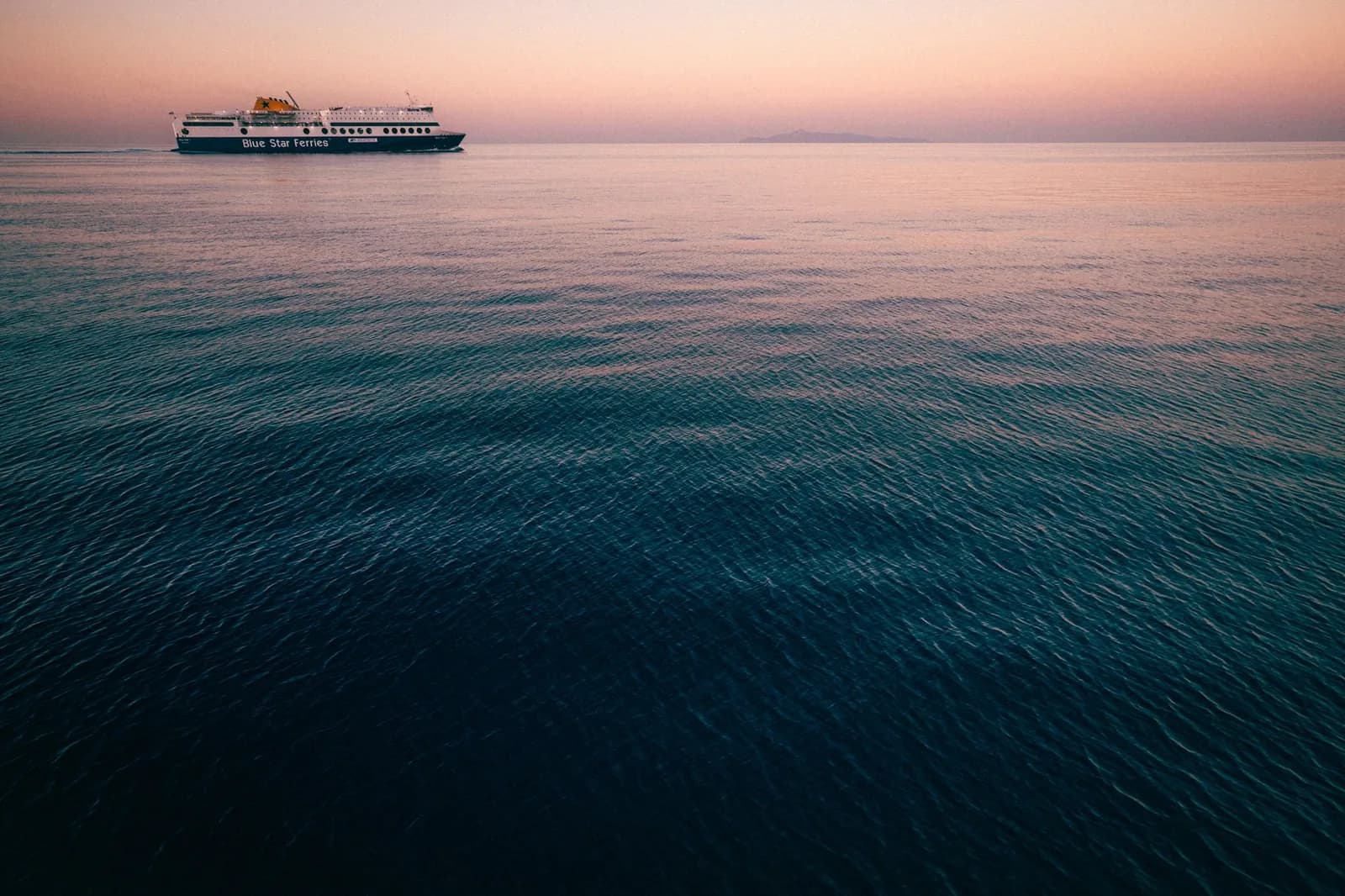 A Blue Star Ferries ship sailing across a calm sea at dusk under a pink sky, with a distant island on the horizon