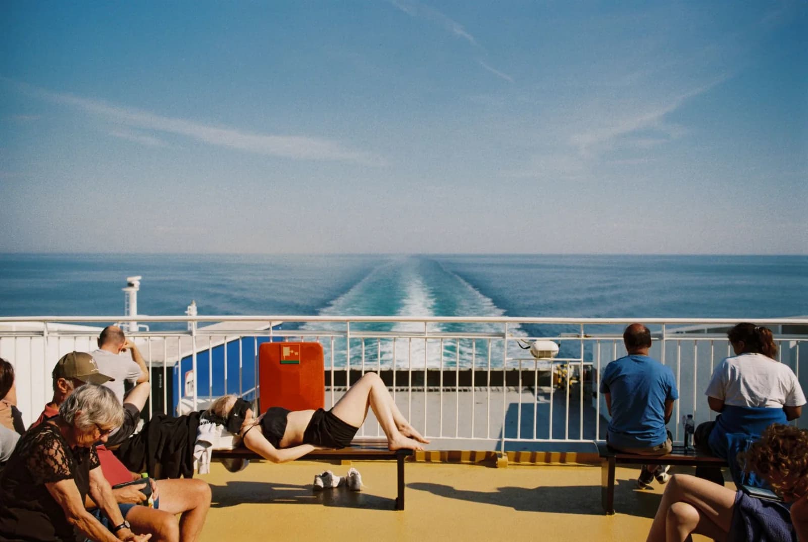 Passengers sitting on the sunny open deck of a large ferry, with the wake trailing across a calm blue sea behind them