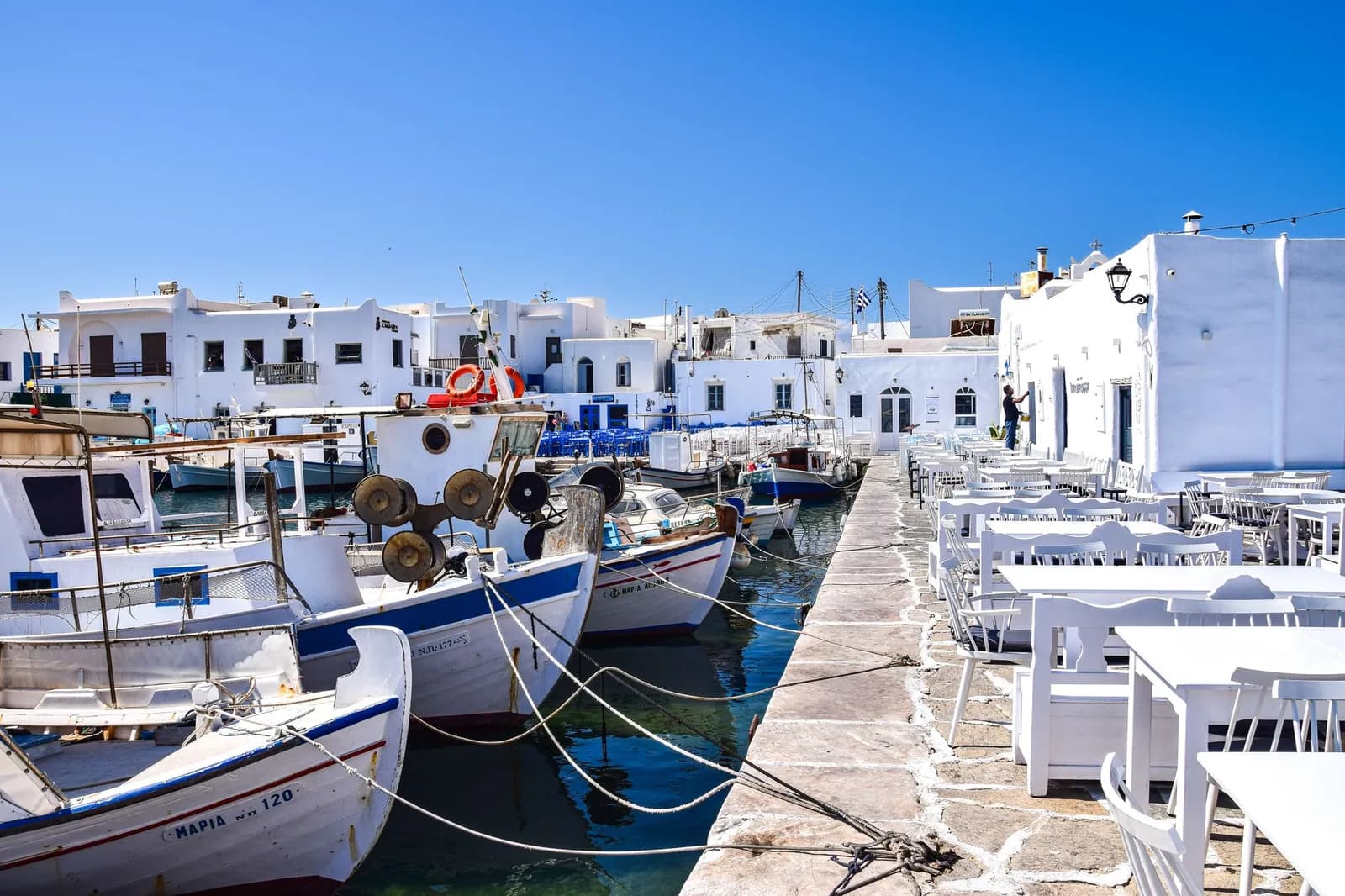 Fishing boats moored along the quay of Naoussa harbour on Paros, with taverna tables set out beside them