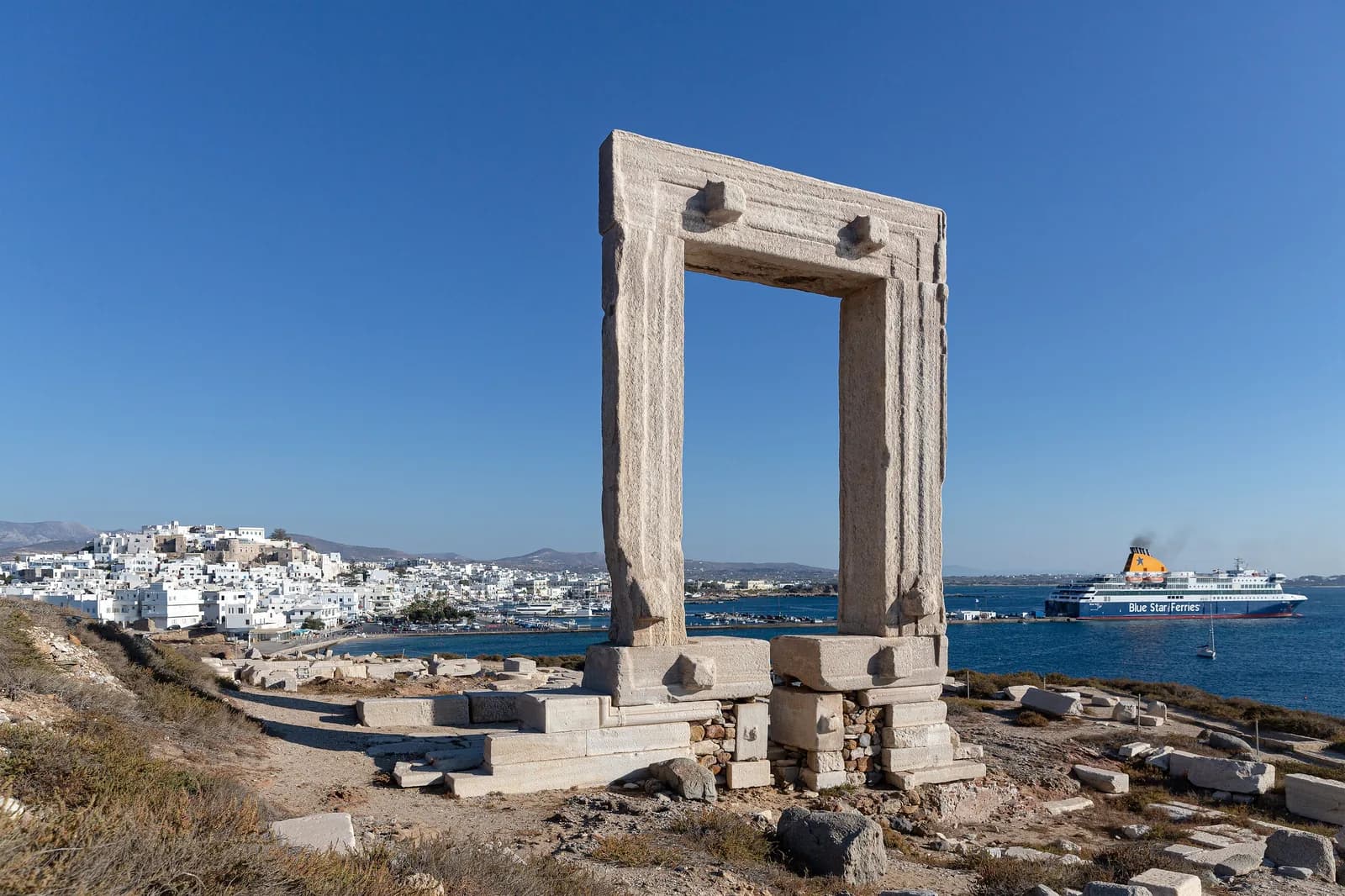 The Portara marble gateway on Palatia with the white houses of Naxos Chora and a Blue Star ferry docked in the port behind it