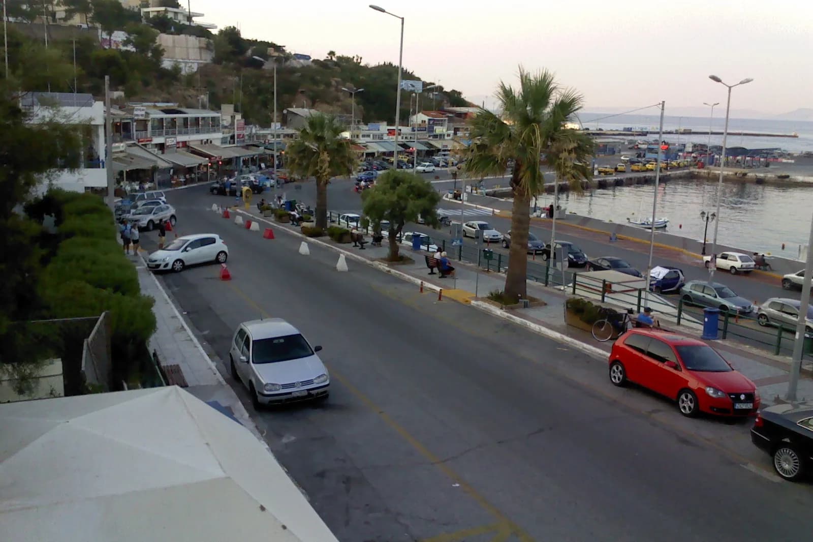 Waterfront street in Rafina, Greece, with taverna awnings on the left, palm trees and the harbor on the right