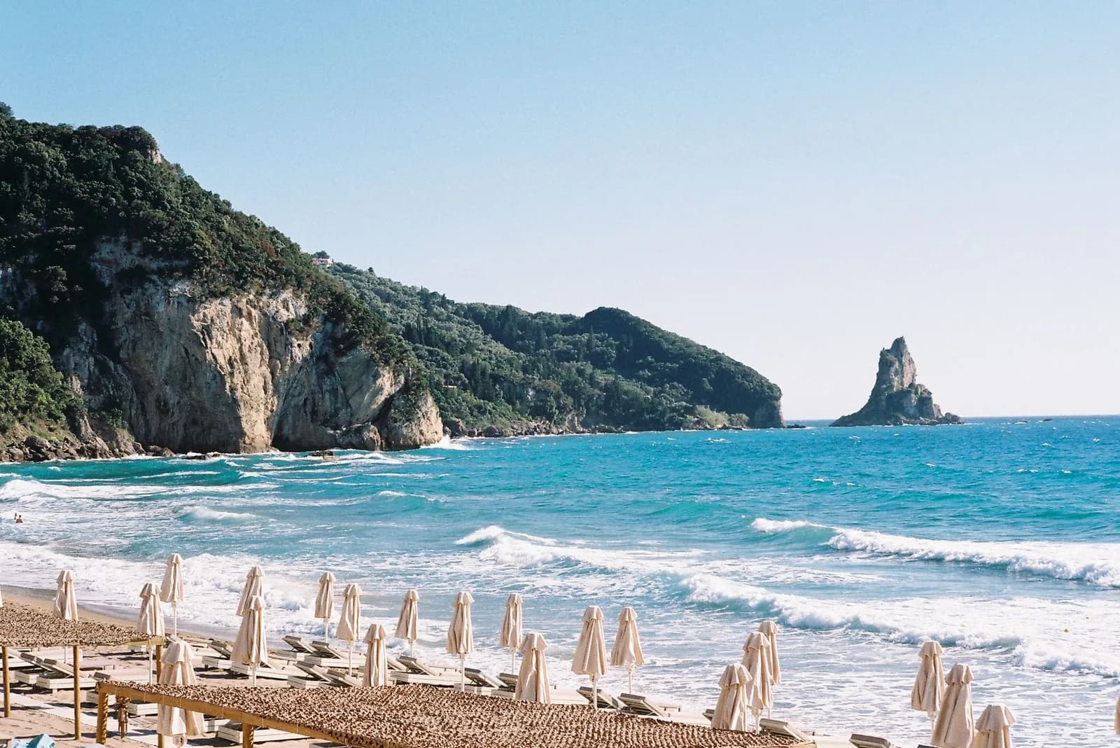 Agios Gordios beach on the west coast of Corfu, rows of closed parasols on the sand, waves coming in, wooded cliffs on the left and the Ortholithos rock stack standing in the sea
