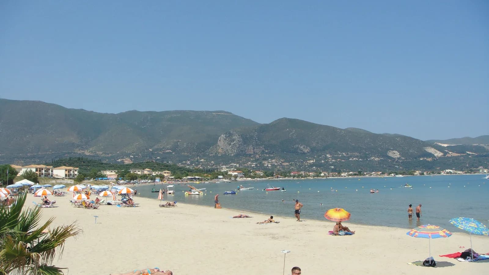 Long sandy beach at Alykes on the north-east coast of Zakynthos, with parasols along calm shallow water and the island's mountains behind the bay