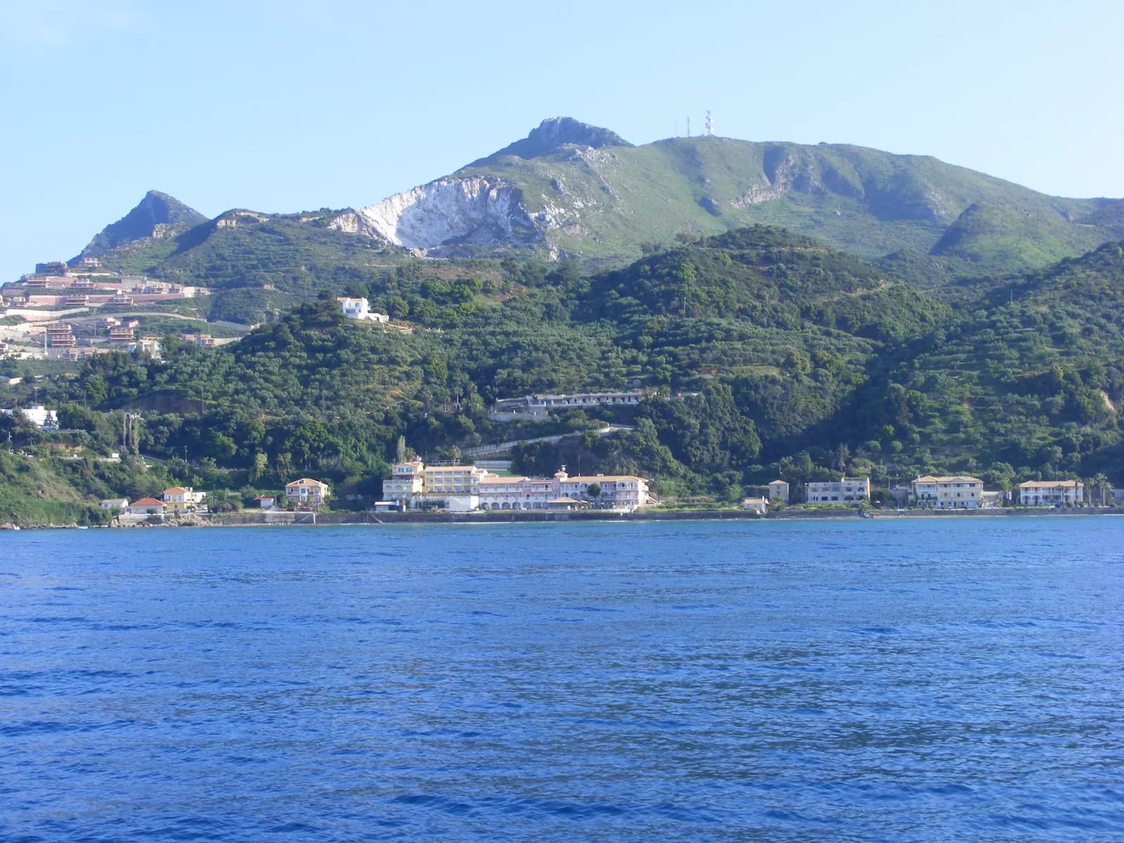 Argassi's seafront hotels seen from the sea, strung along the shore below the green slopes and quarry-scarred peak of Mount Skopos, Zakynthos