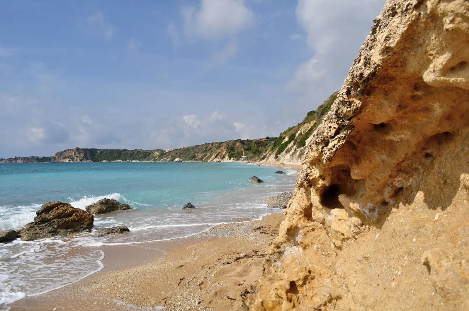 Avithos beach on the south coast of Kefalonia, sand and shingle under low ochre cliffs with clear turquoise water and a few villas on the headland beyond