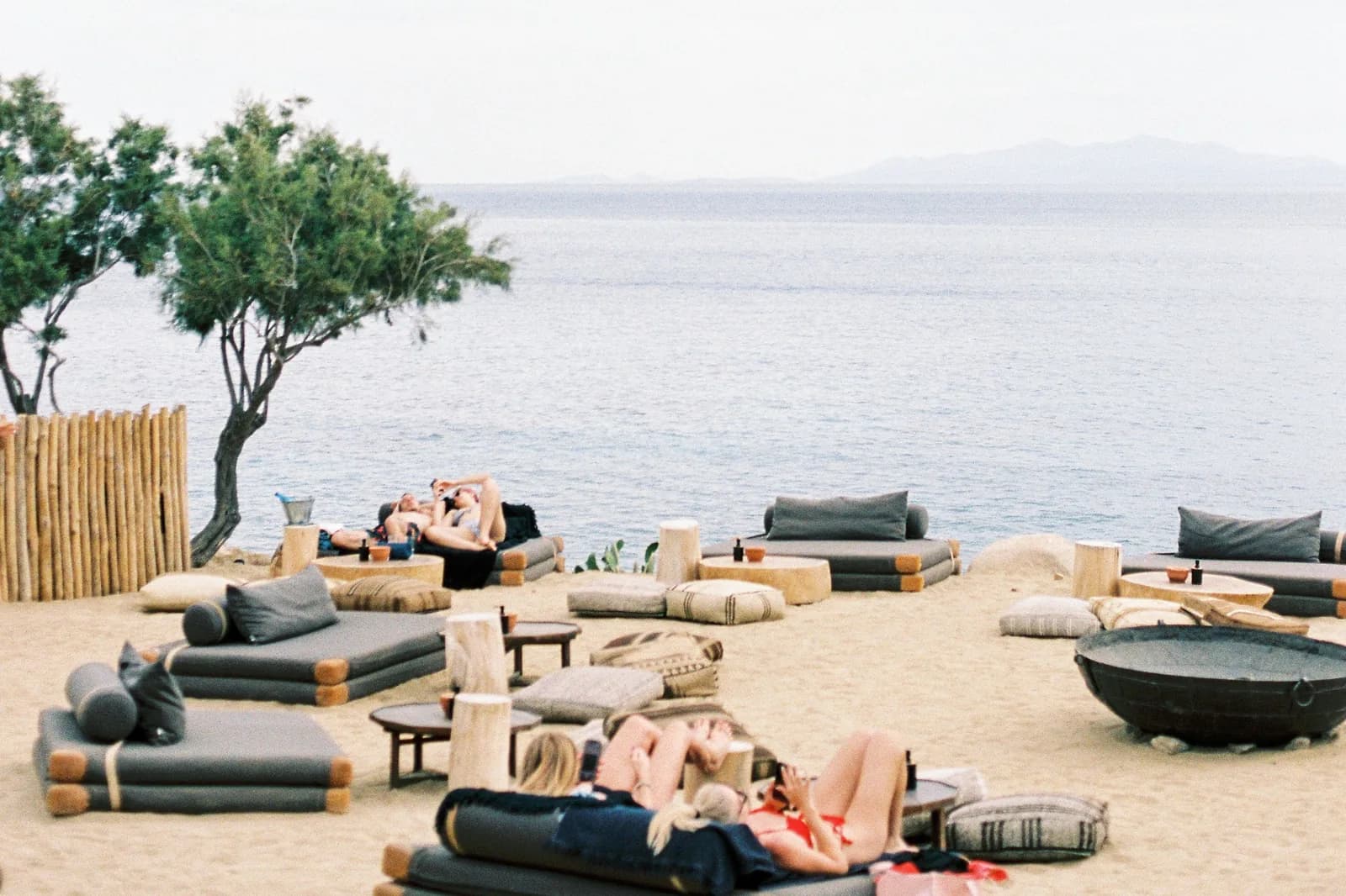 Low daybeds and cushions arranged on the sand of a Mykonos beach club under a wind-shaped tree, with the sea and the outline of another island behind