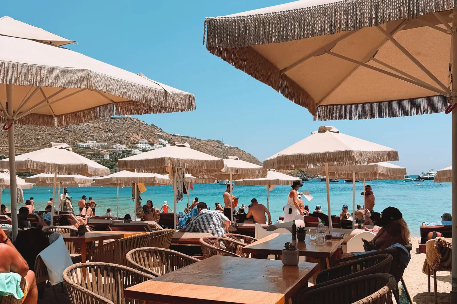 Fringed parasols and restaurant tables set out on the sand of an organised Mykonos beach, with turquoise water and a dry hillside of white houses behind