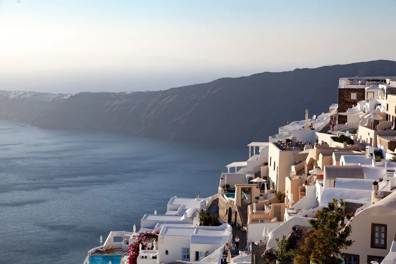 Hotels stacked down the caldera cliff on Santorini at sunset, terraces and small plunge pools cut into the rock above the water