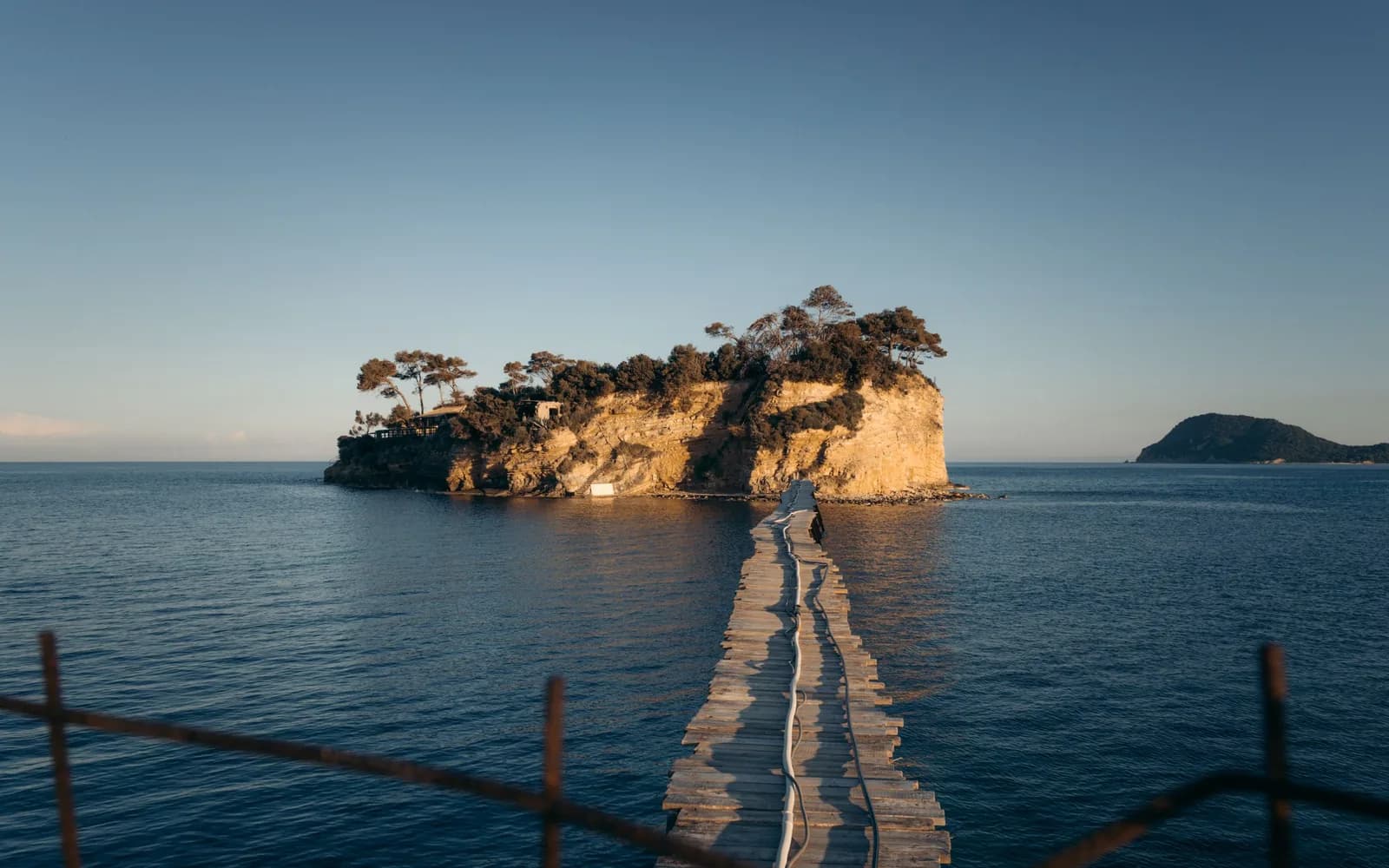 Wooden footbridge leading across calm water to pine-covered Cameo island at Agios Sostis, at the west end of Laganas Bay, Zakynthos, in low evening light