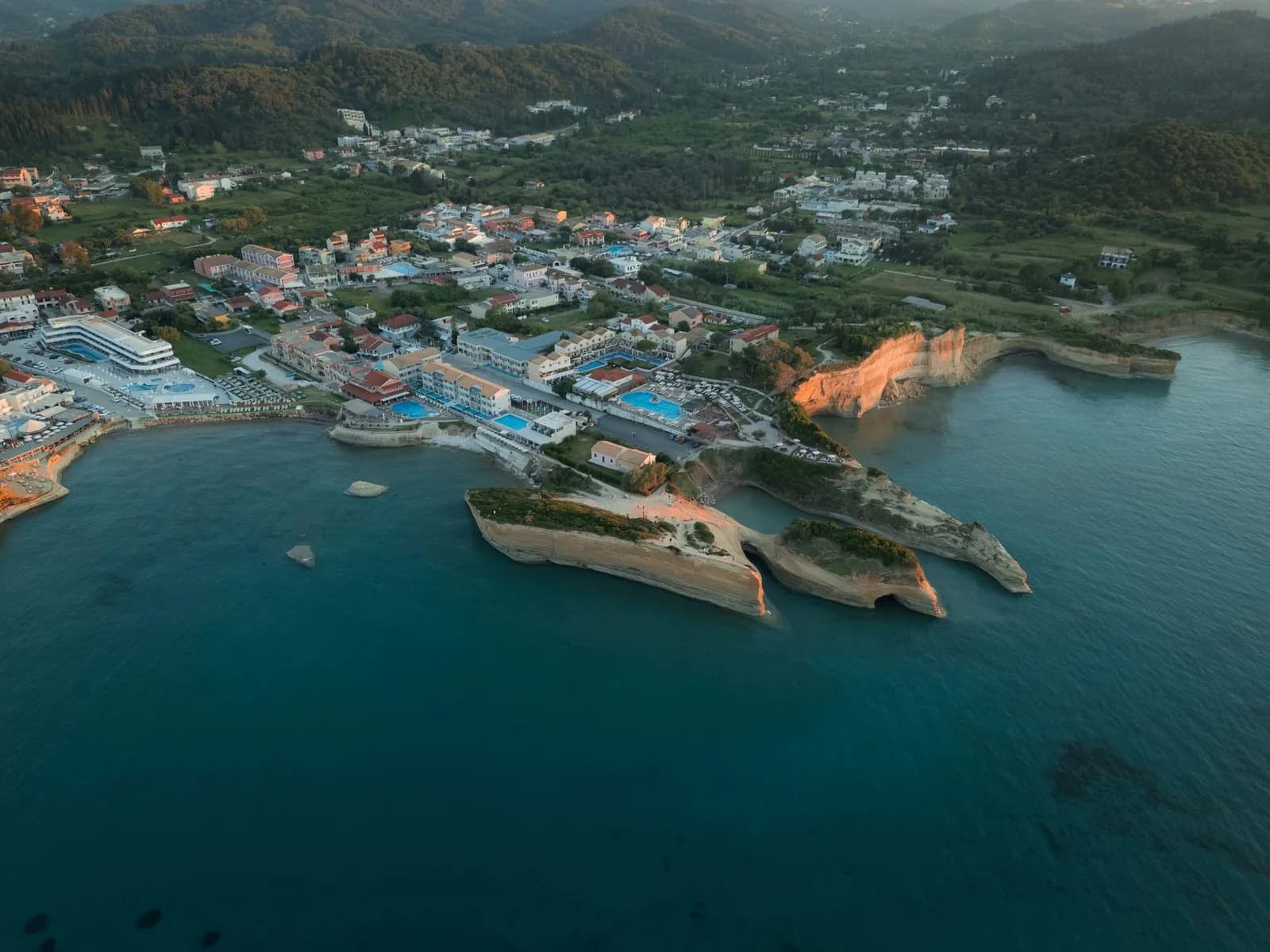 Aerial view of Sidari on the north coast of Corfu at dusk, the sandstone fingers of the Canal d'Amour reaching into the sea in front of hotels with pools and the green hills behind