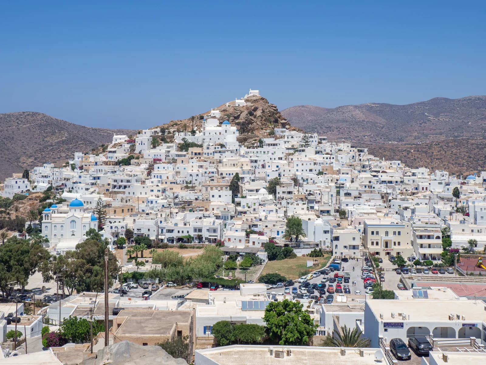 Chora, the main village of Ios, a hill of white cube houses rising to a rocky summit with a small white chapel, the blue-domed cathedral on the left and bare brown hills behind