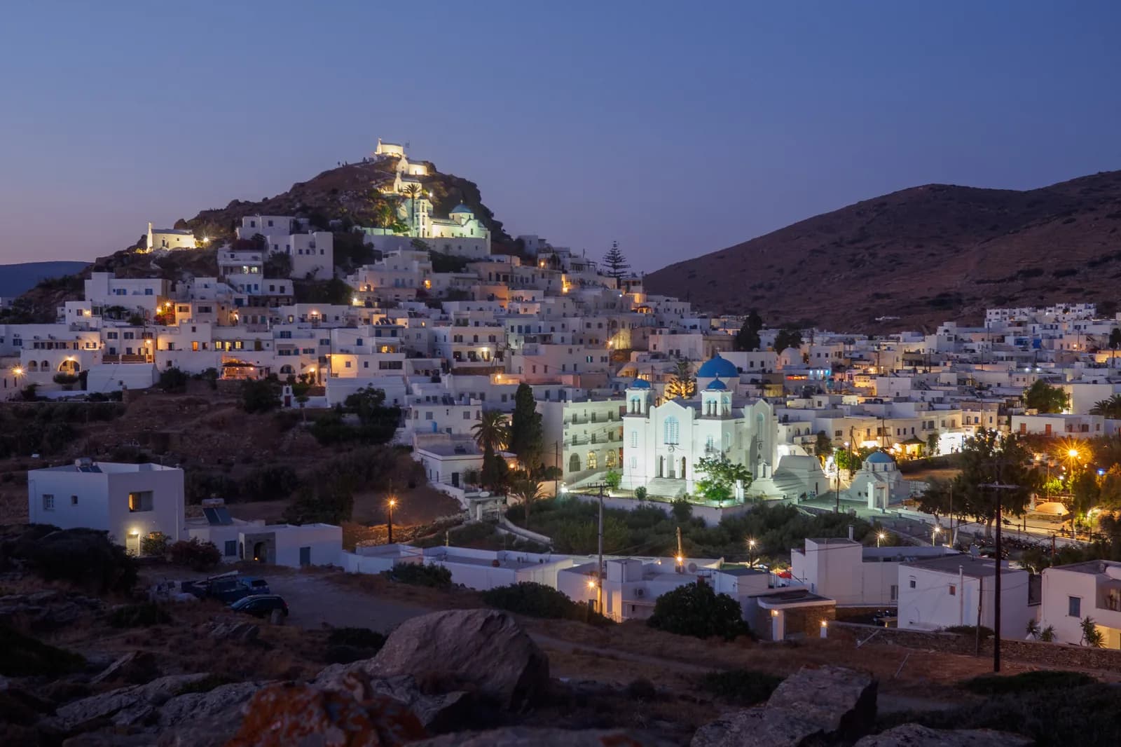 Chora, the hilltop village of Ios, at dusk, with the white houses climbing to the lit chapels on the summit and the floodlit cathedral with blue domes below