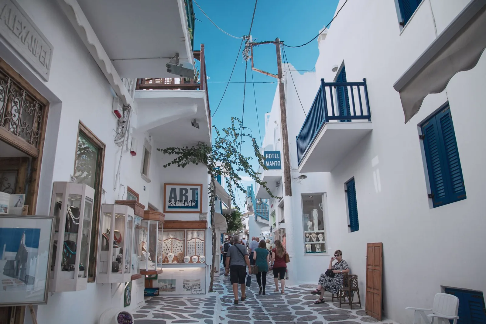 A narrow whitewashed lane in the old town of Mykonos Chora, with jewellery shop windows, blue shutters and a hotel sign hanging above the paving