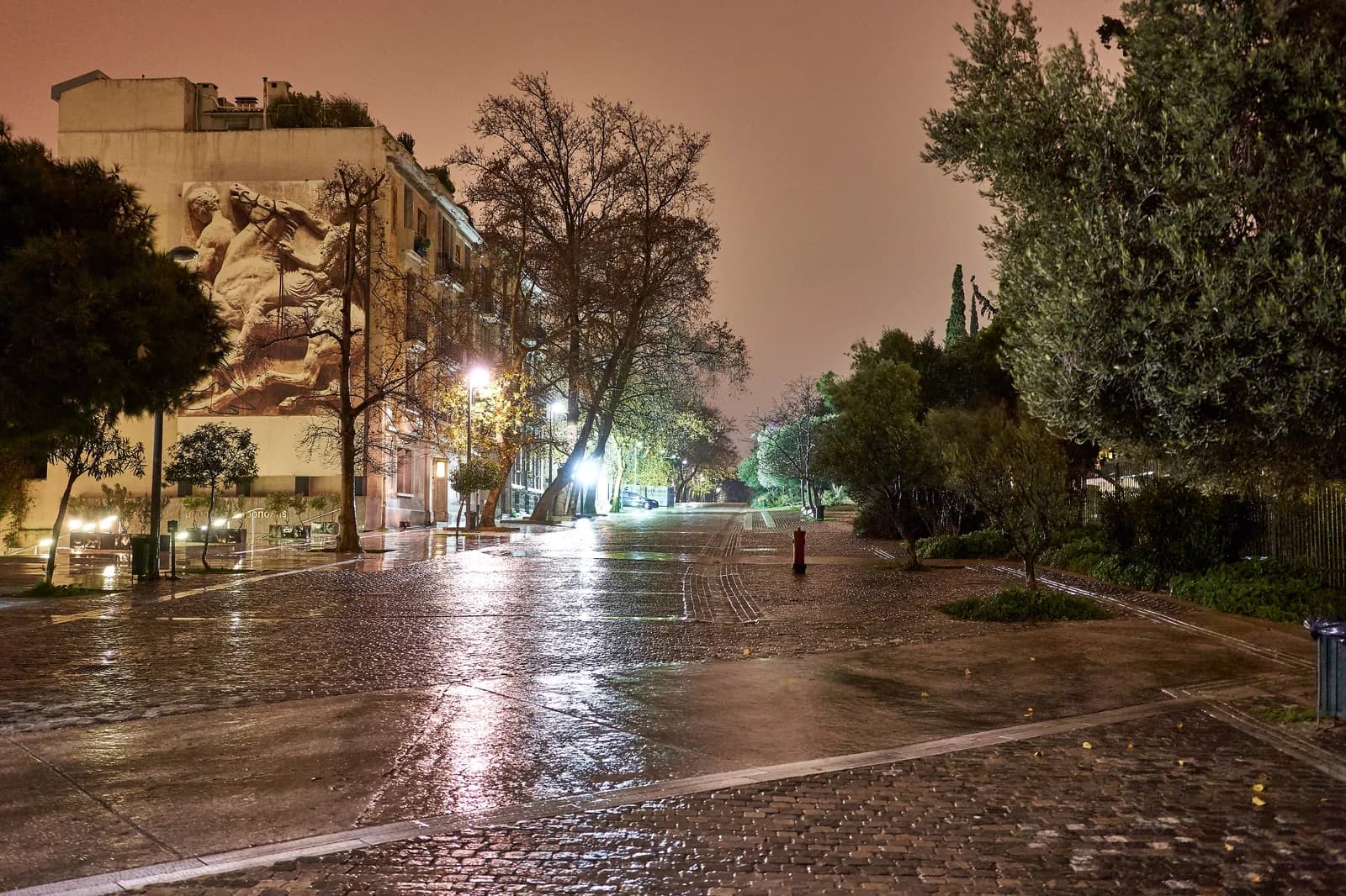 The cobbled Dionysiou Areopagitou pedestrian street in Makrygianni, Athens, wet at night, with a large mural on a building near the Acropolis Museum