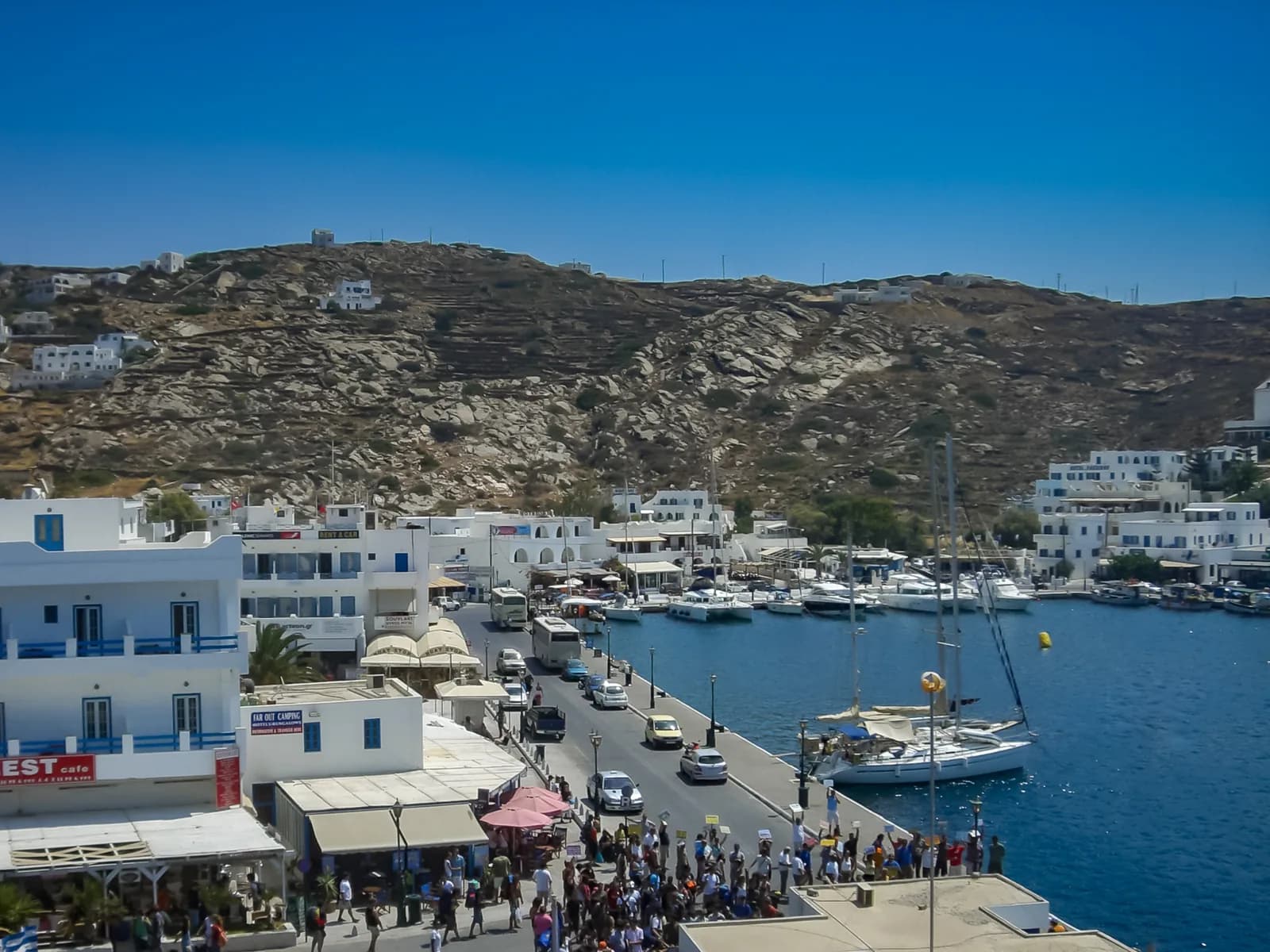 The harbour of Ios at Gialos, with white hotels along the quay, yachts moored in the blue water, buses on the harbour road and a crowd waiting at the ferry dock below a rocky hillside