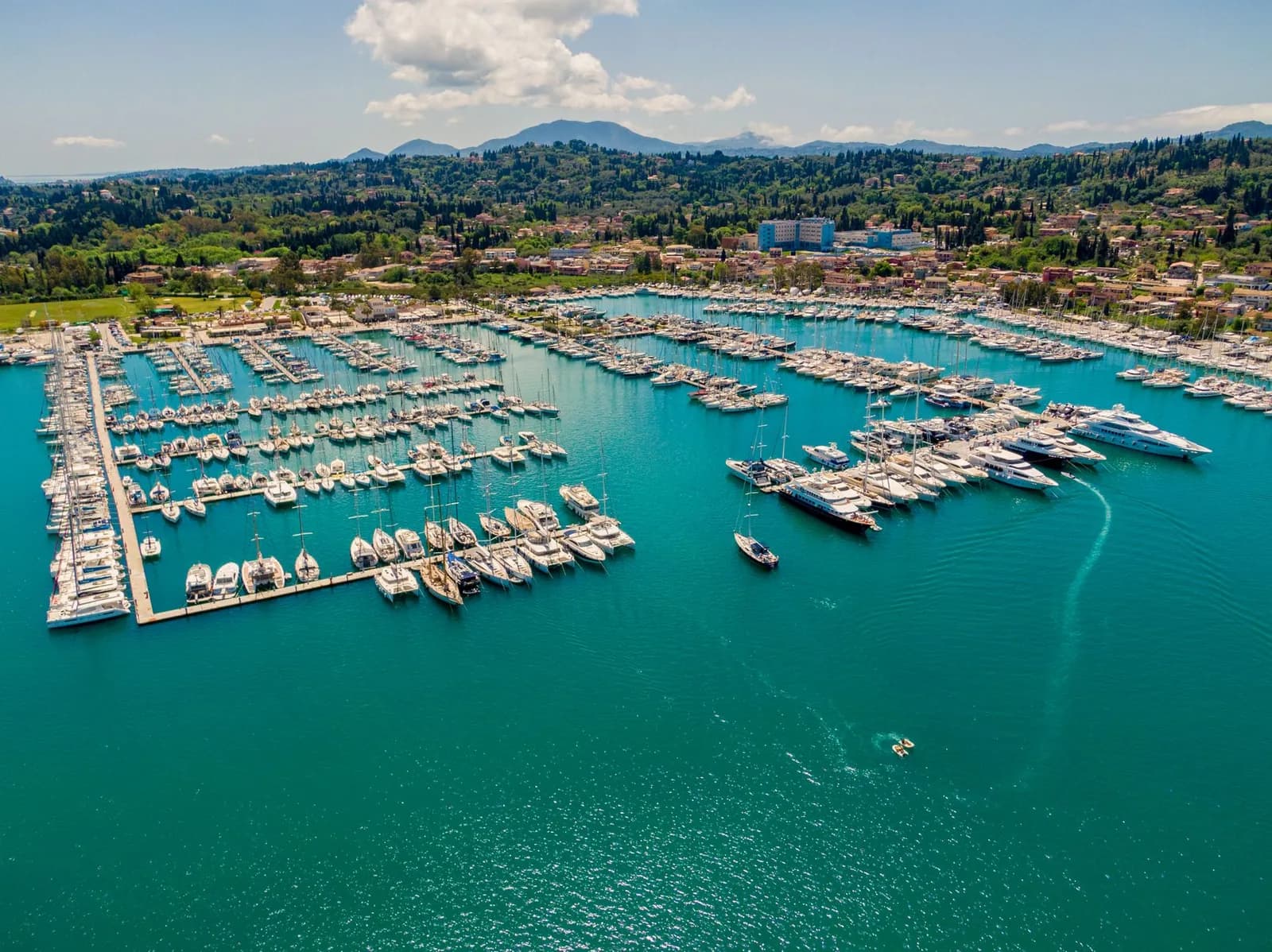 Aerial view of the marina at Gouvia on Corfu, hundreds of yachts moored along long pontoons in turquoise water, with the village and wooded hills rising behind