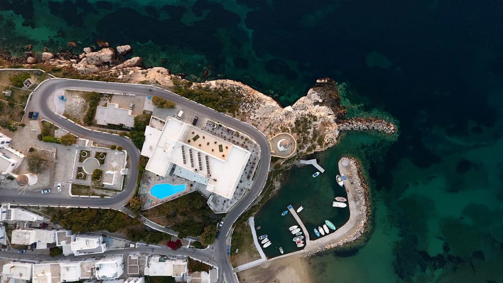 A hotel and its pool on the headland above a small fishing harbour on the coast of Paros