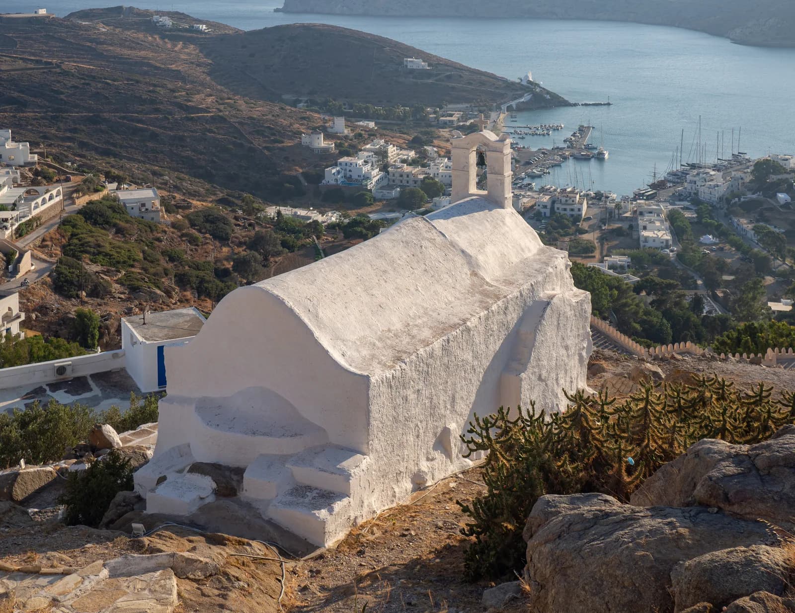 A whitewashed chapel on the hill of Chora, Ios, looking down over houses to the harbour of Gialos, the moored yachts and the long bay stretching west towards Koumbara