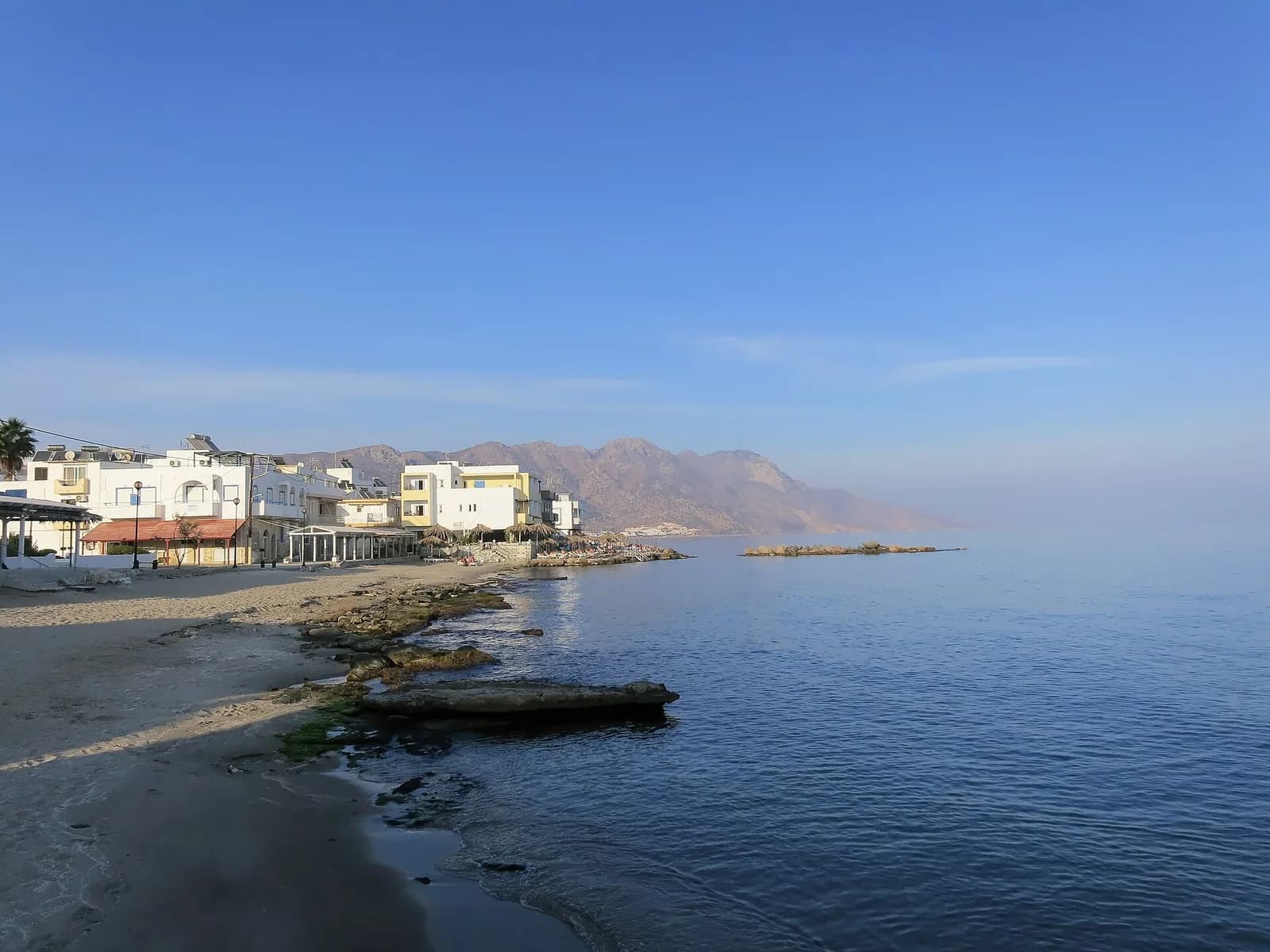 The waterfront at Kardamena on the south coast of Kos, with white buildings and tavernas along the beach and mountains in the distance