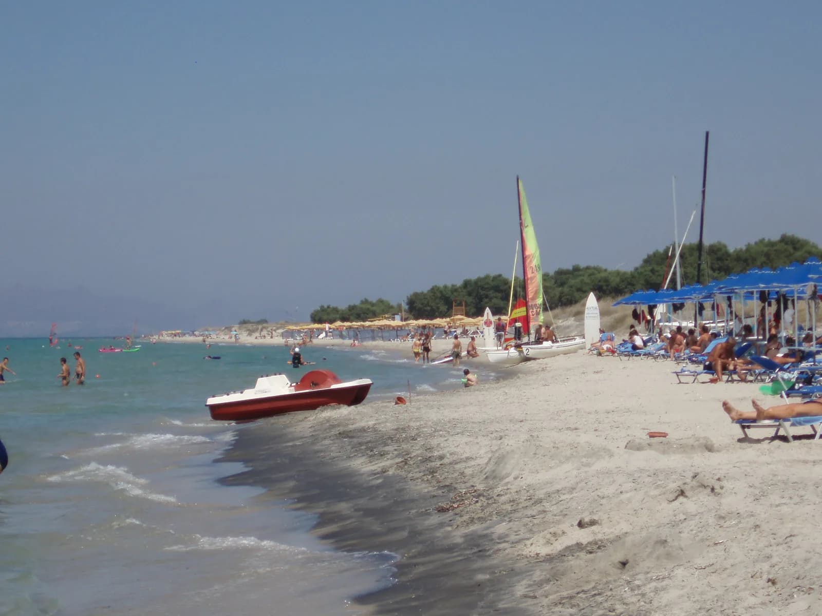 The long sandy beach at Marmari on the north coast of Kos, with swimmers in shallow water, parasols and small sailing boats pulled up on the sand