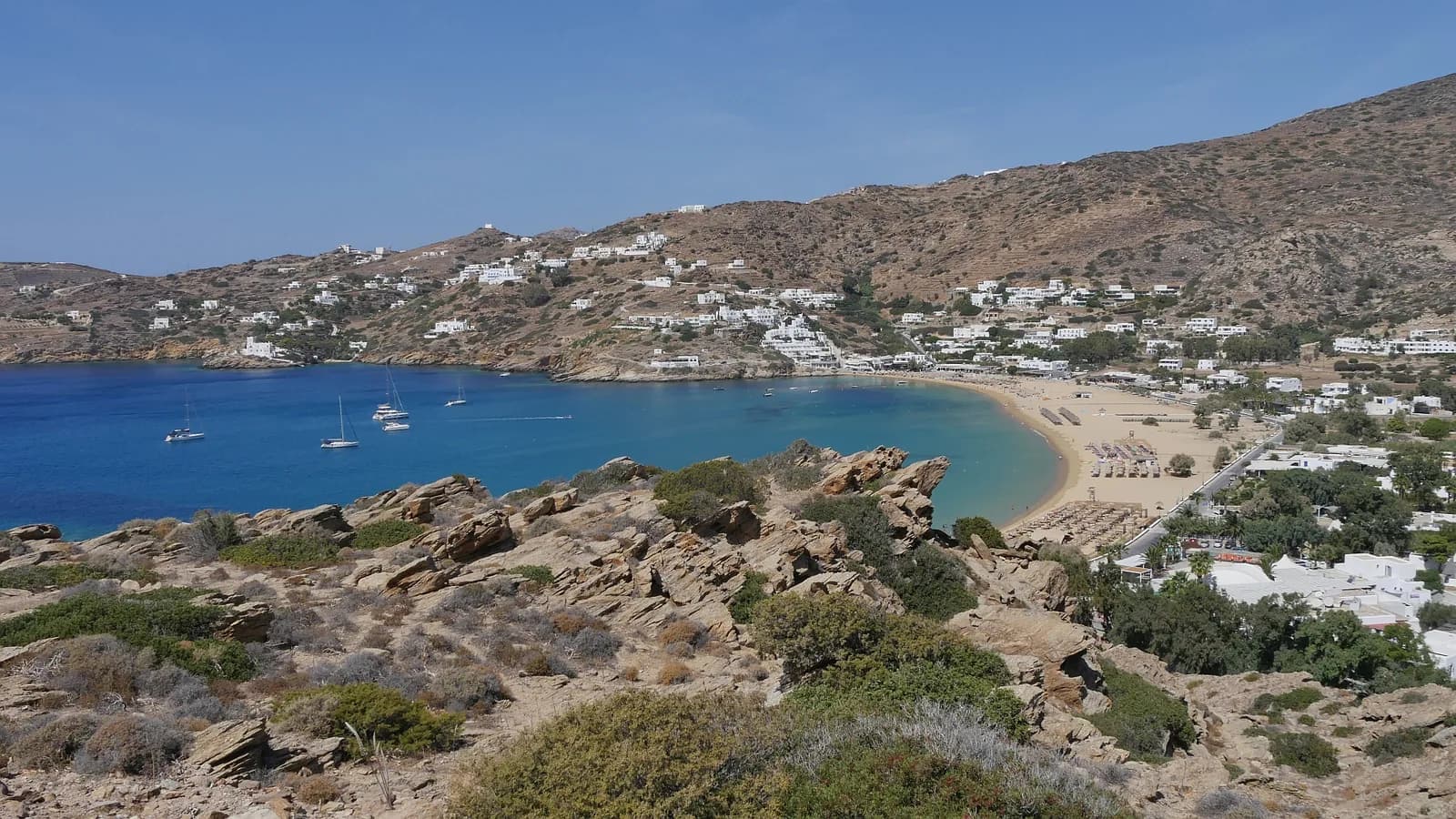Mylopotas bay on Ios seen from the rocky headland: a long curve of golden sand, hotels spread up the hillside and yachts anchored in deep blue water