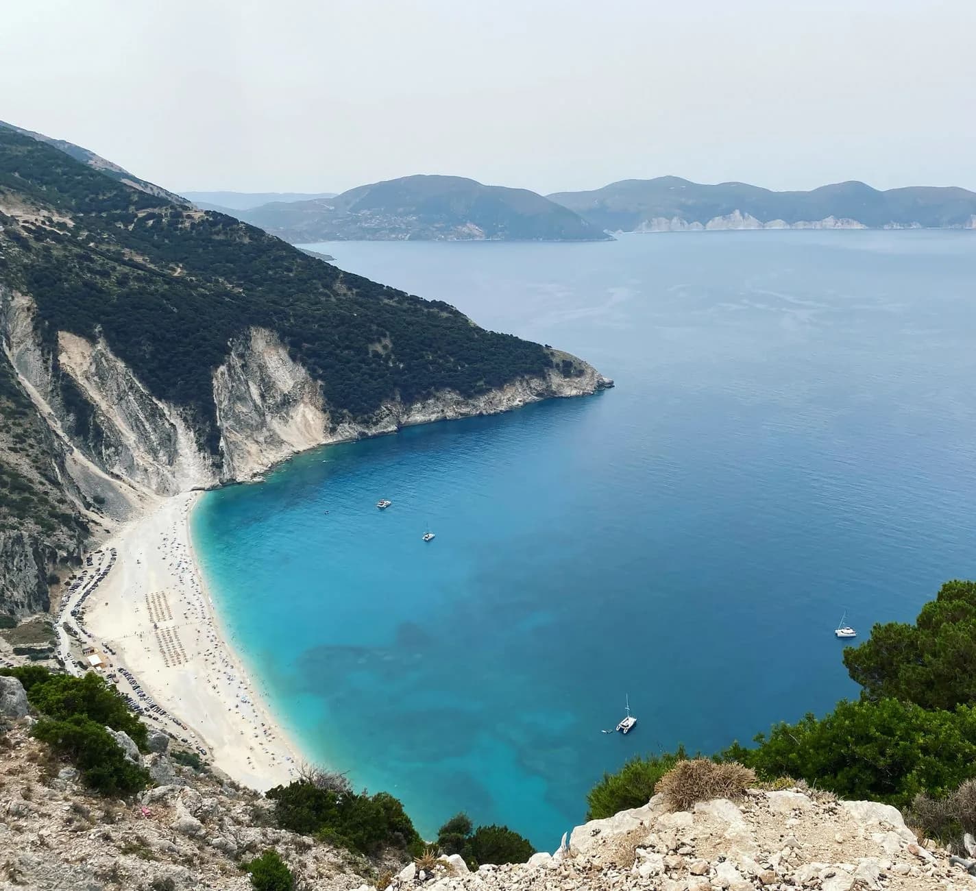Myrtos beach on Kefalonia seen from the cliff road above, a white shingle crescent under limestone cliffs with turquoise shallows, catamarans anchored offshore and the headlands of the north coast beyond