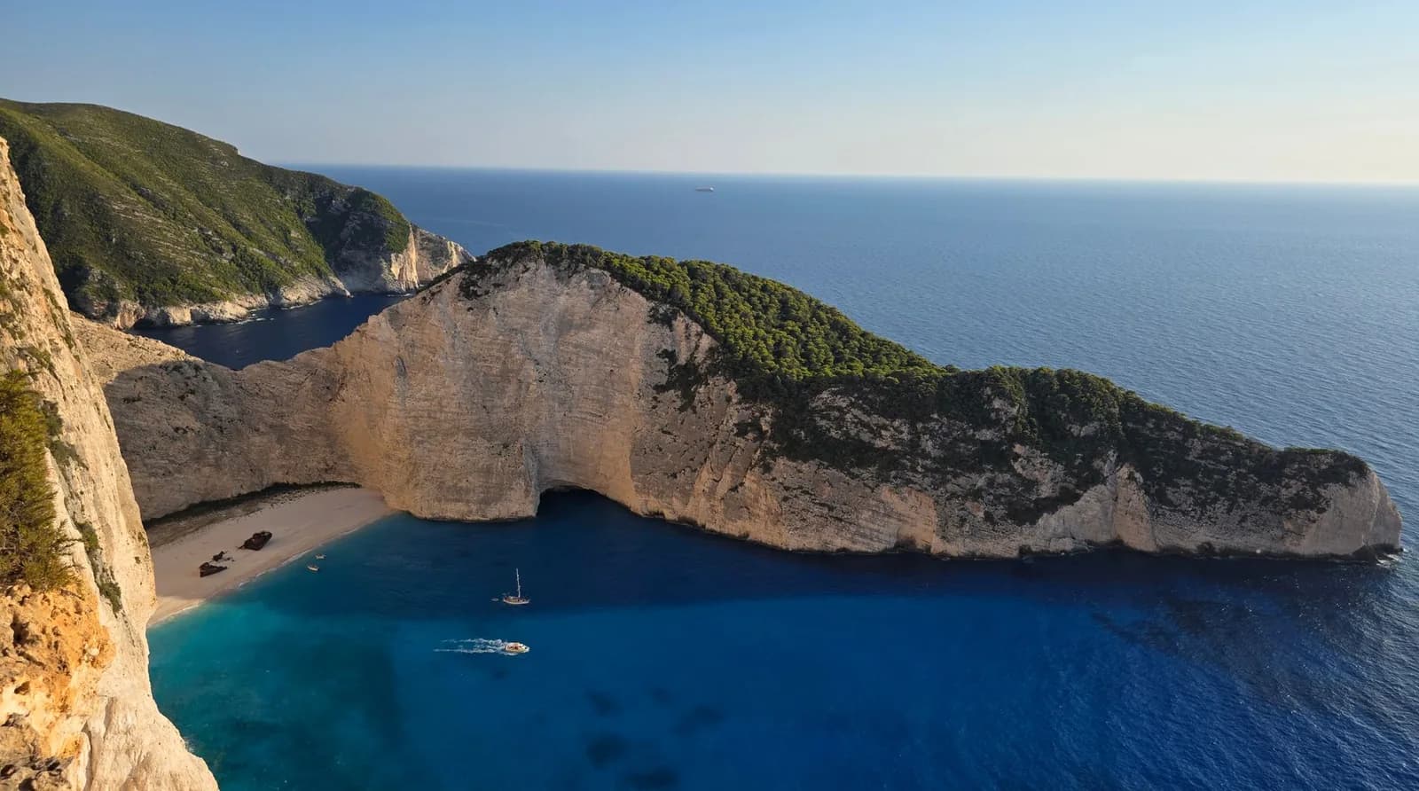 Navagio, the Shipwreck beach on Zakynthos, seen from the clifftop viewpoint: a white sand cove with the rusting wreck below limestone cliffs topped with pine, above deep blue sea