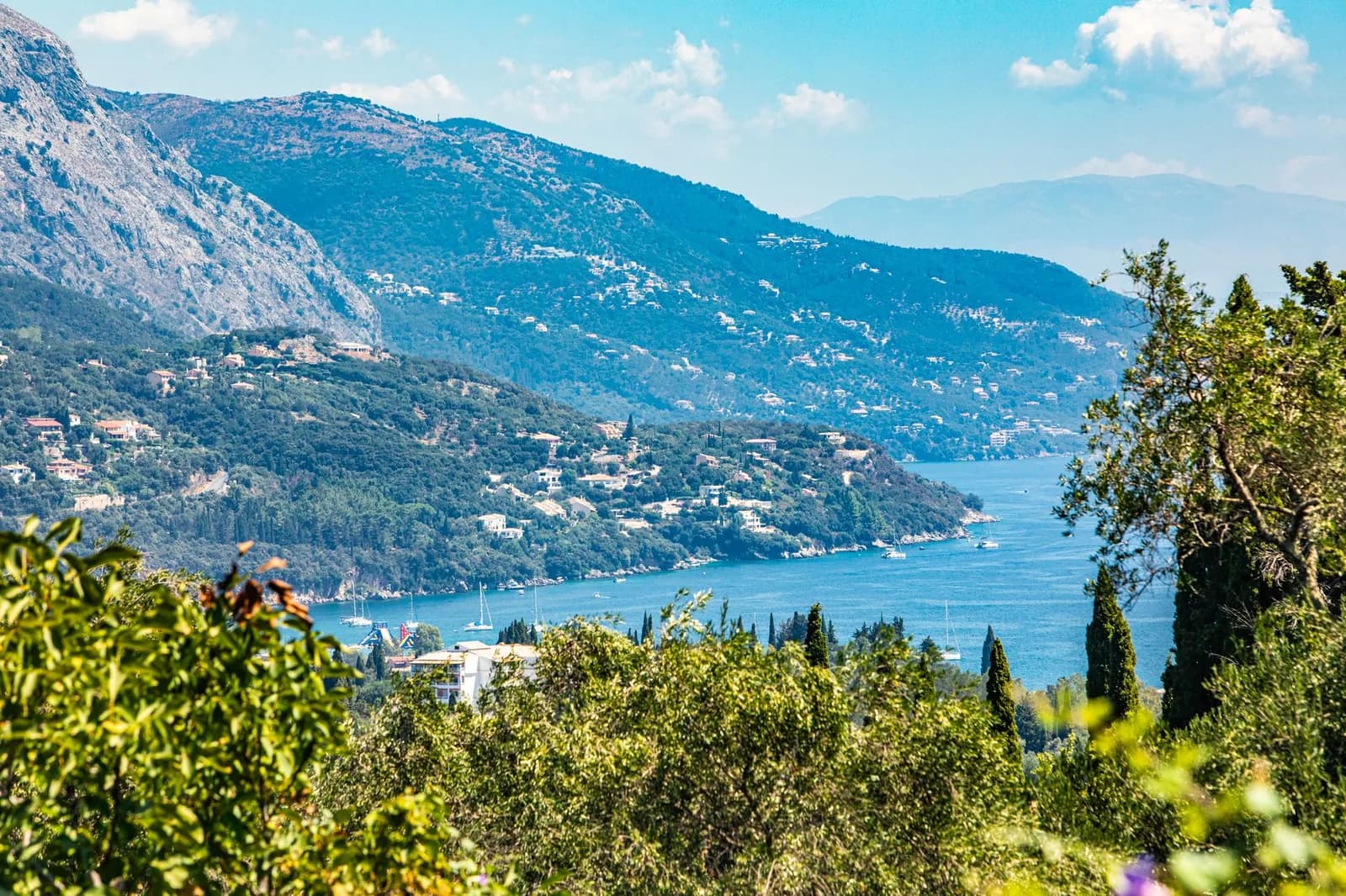 The north-east coast of Corfu seen from the hills, villas scattered across the green slopes of Mount Pantokrator above a narrow channel of blue water with yachts at anchor and the mainland mountains beyond