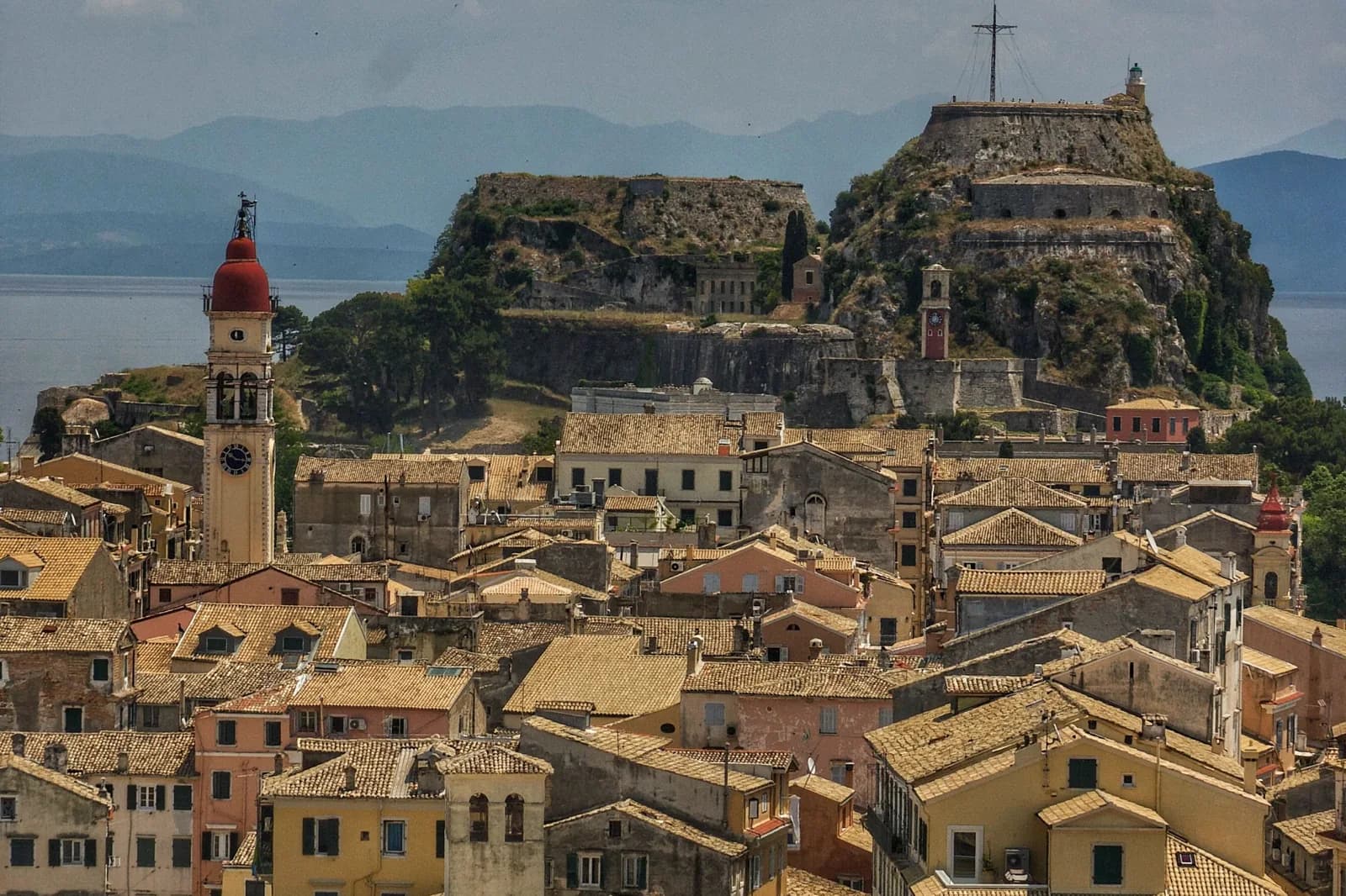 The tiled rooftops of Corfu old town with the red-domed bell tower of Saint Spyridon on the left and the two peaks of the Old Fortress behind, with the mainland mountains across the water