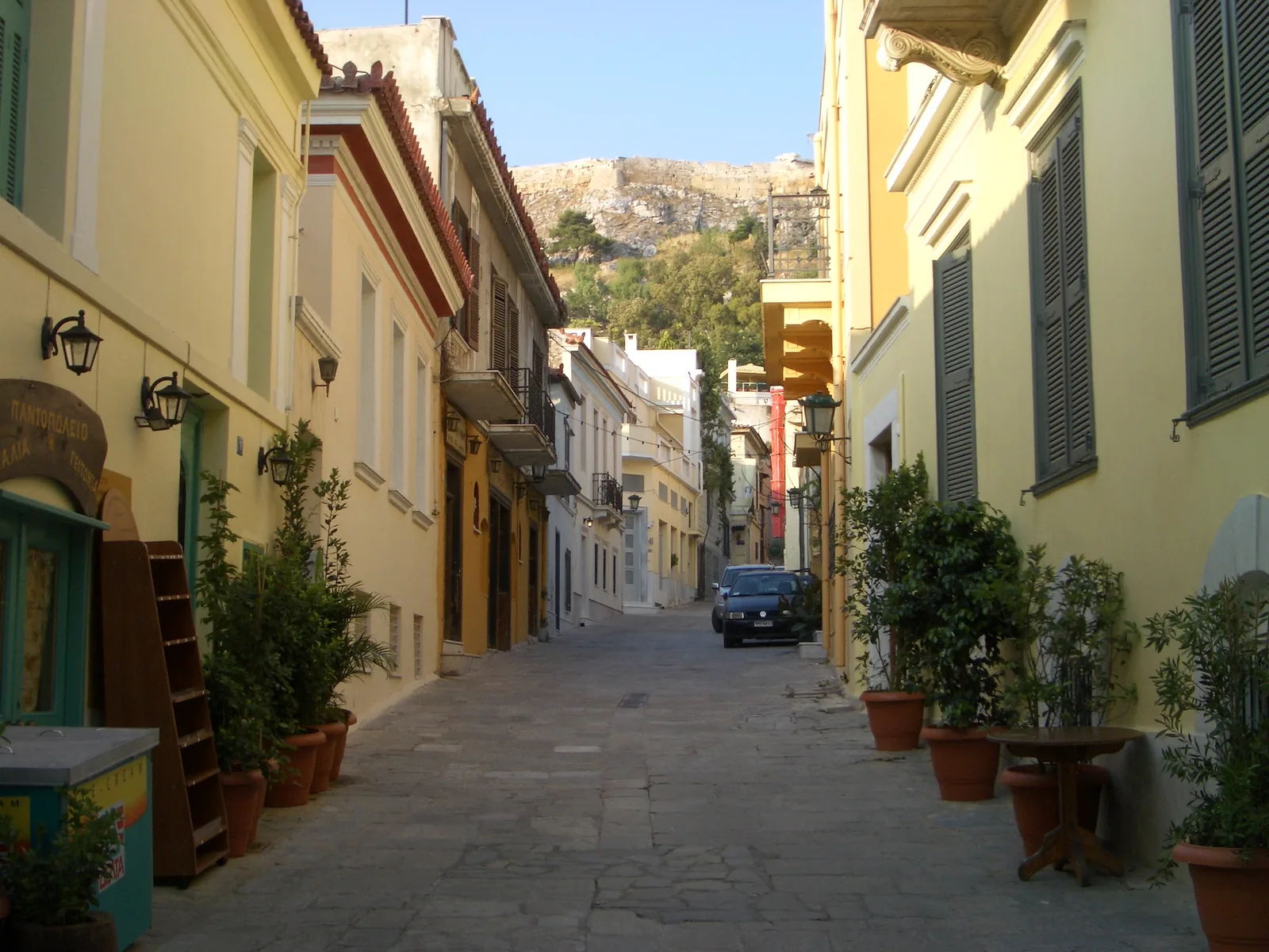 A quiet cobbled lane in Plaka, Athens, lined with yellow neoclassical houses and potted plants, with the rock of the Acropolis rising behind