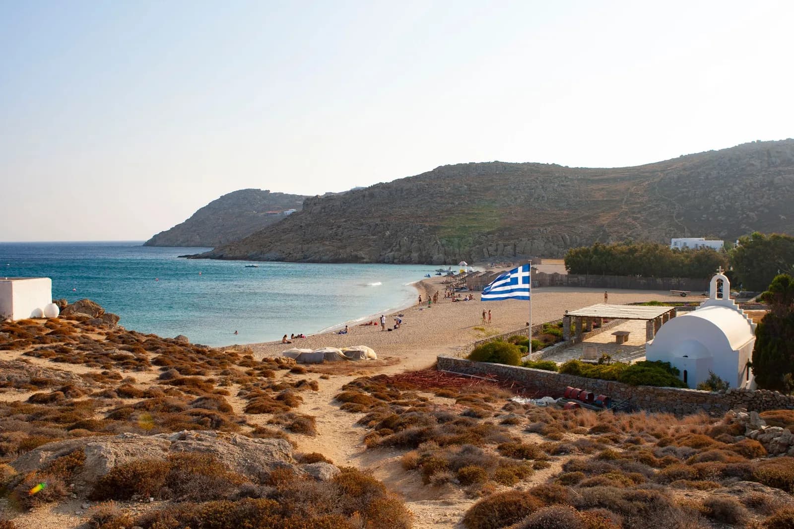 A wide sandy bay on the far side of Mykonos with a small whitewashed chapel and a Greek flag above it, dry hills running down to calm water and few people on the sand