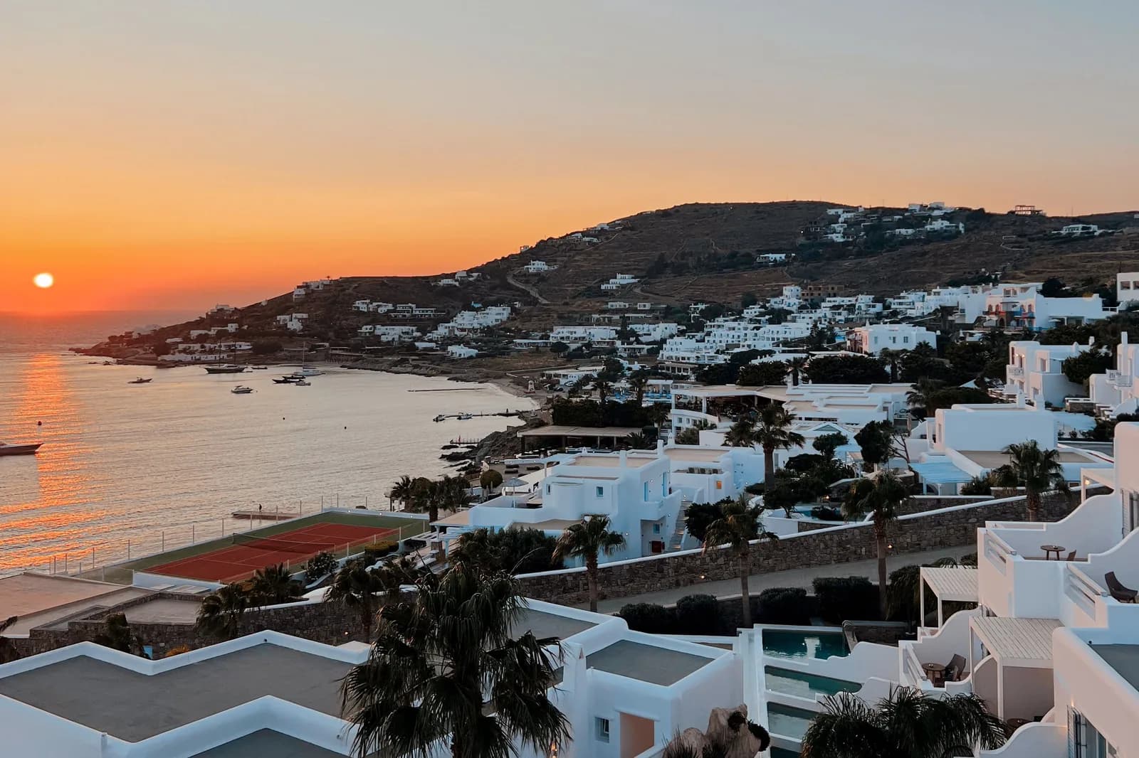 Terraced white hotel buildings with small pools and palm trees running down to a bay on Mykonos at sunset, with more hotels spread across the hillside opposite