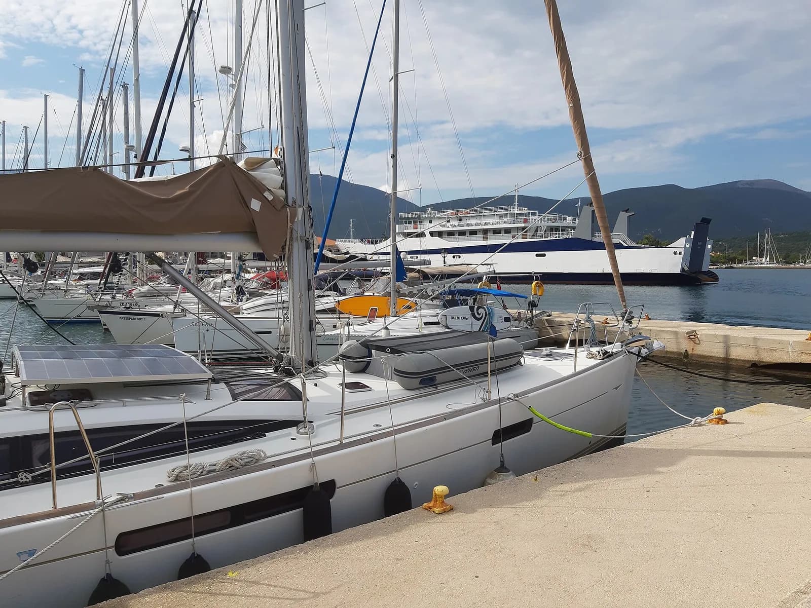 The harbour at Sami on Kefalonia, chartered yachts moored along the quay and a white ro-ro ferry berthed across the bay with the green mountains of Ithaca behind
