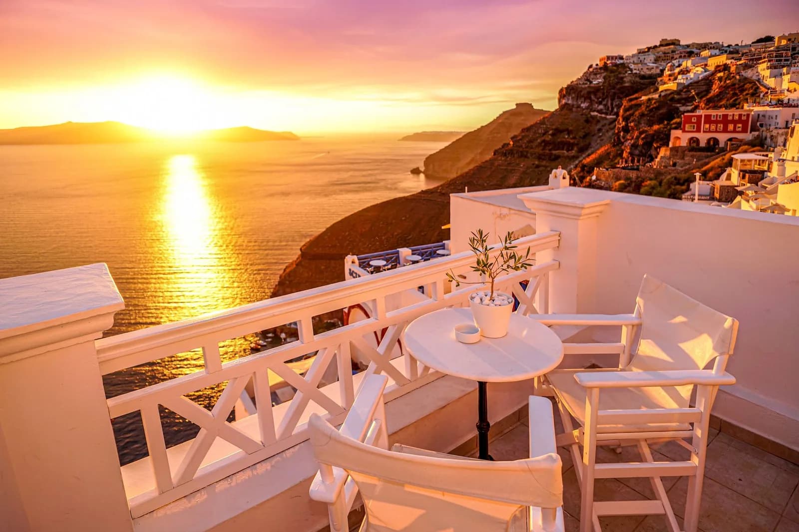A whitewashed hotel terrace on the Santorini caldera at sunset, with the sea and Skaros rock below
