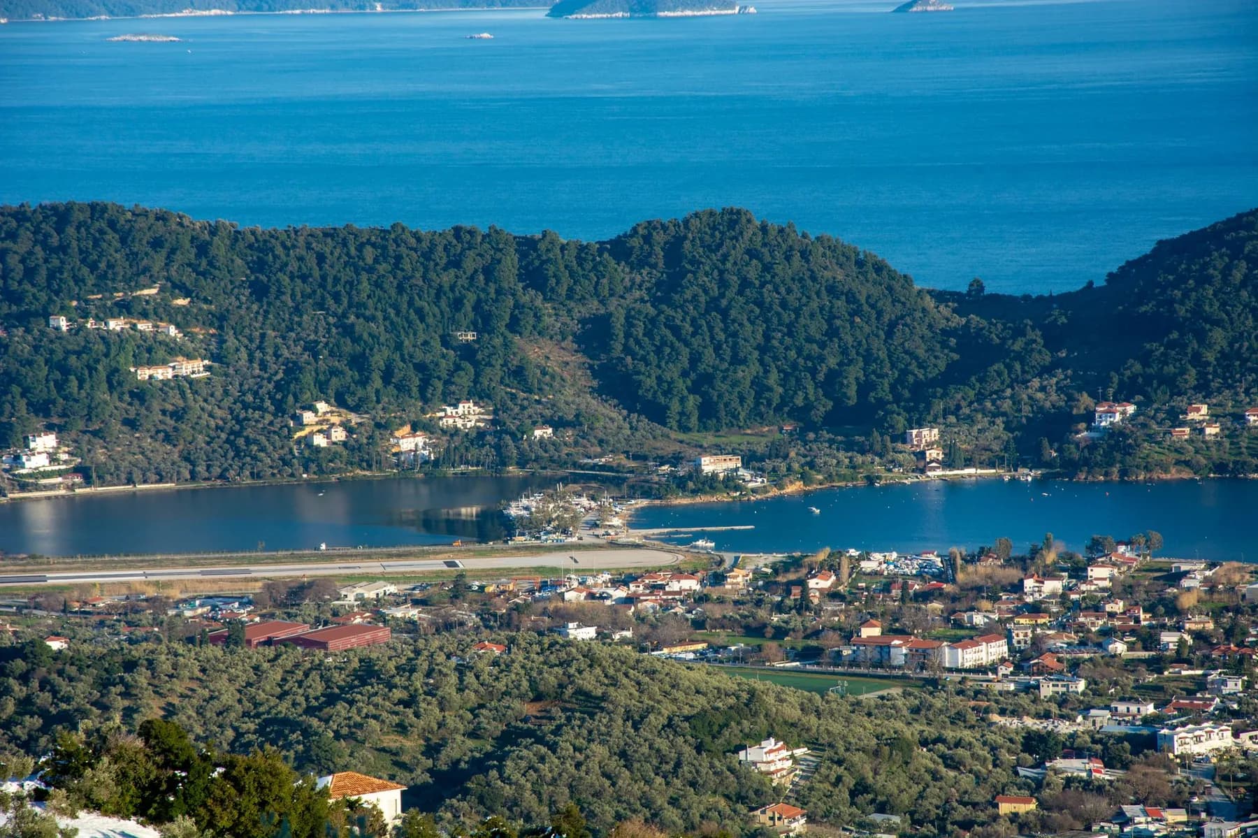 Skiathos seen from the hills above, the airport runway running across a narrow neck of flat land between a lagoon and the bay, with the town spreading south of it and pine-covered hills beyond