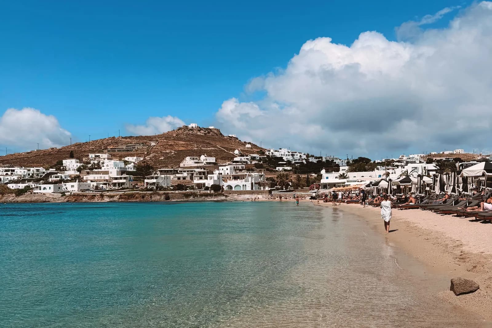 A sheltered south coast bay on Mykonos with rows of sunbeds along the sand, clear shallow turquoise water and white hotels on the dry hillside behind
