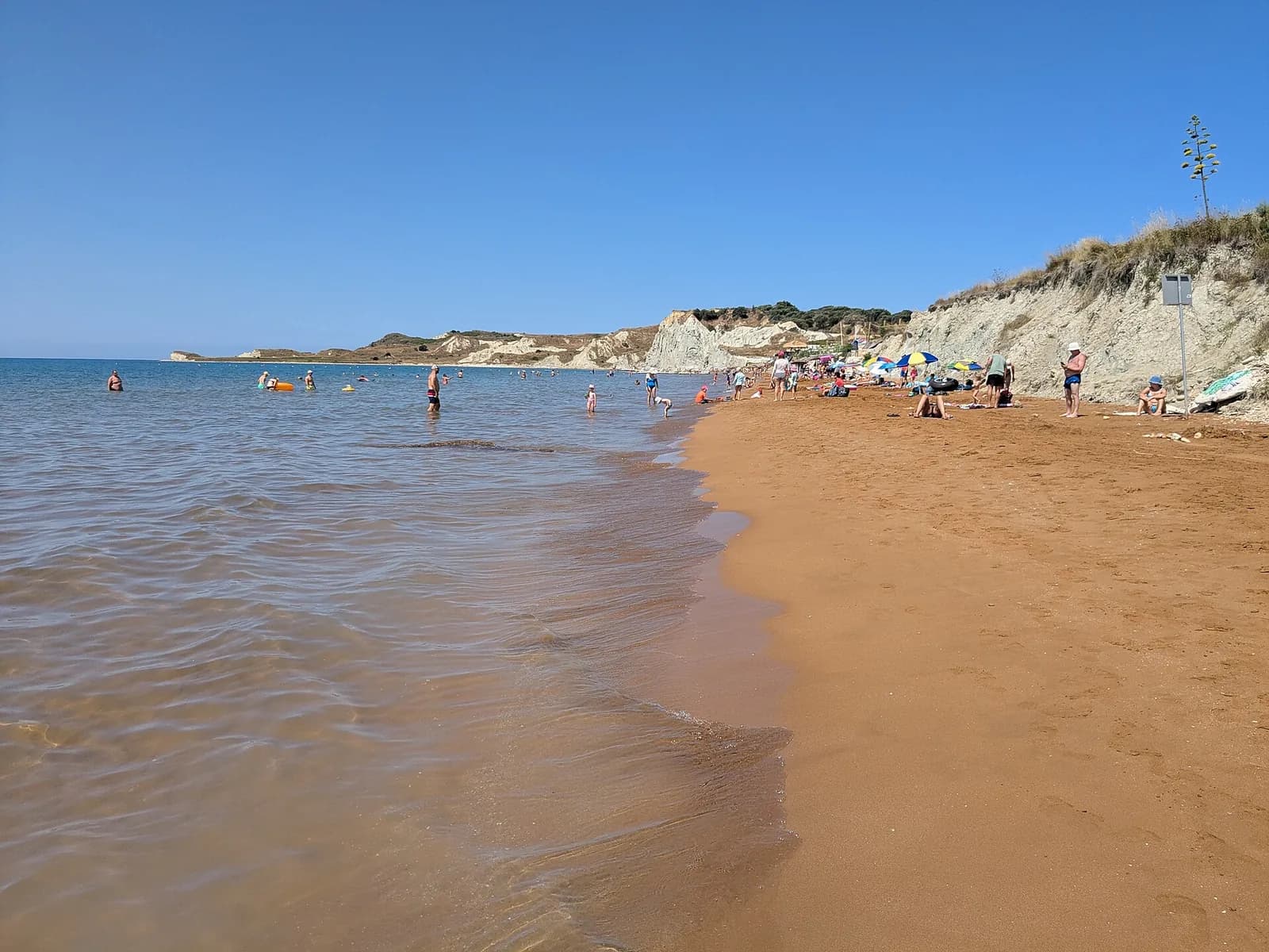 Xi beach on the Paliki peninsula of Kefalonia, orange-red sand running into shallow clear water with white clay cliffs and a line of parasols along the back of the beach